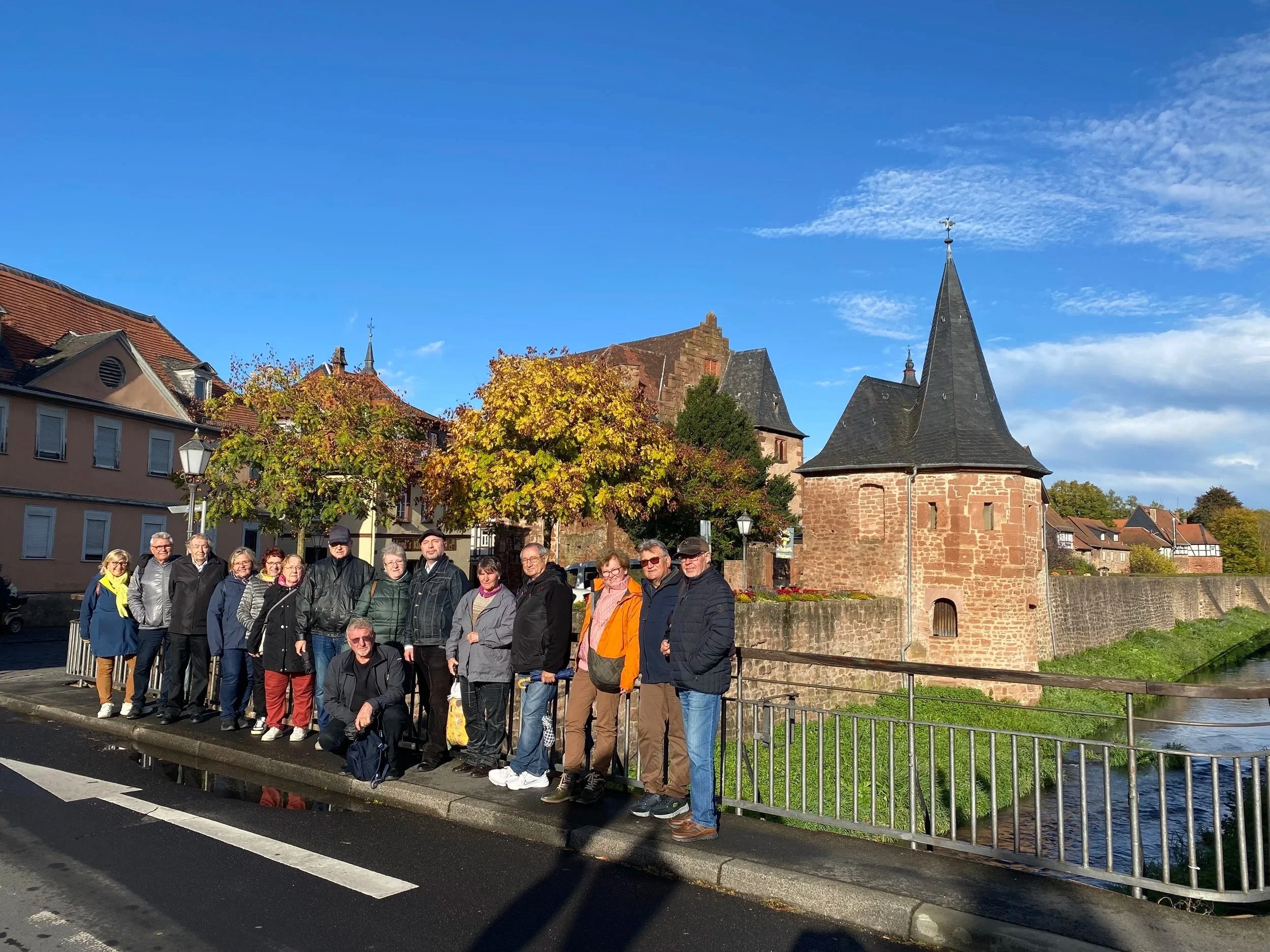 Gruppe von Menschen vor einer alten Stadtmauer und einem Türmchen, herbstliches Laub, blauer Himmel, Stadthaus im Hintergrund.