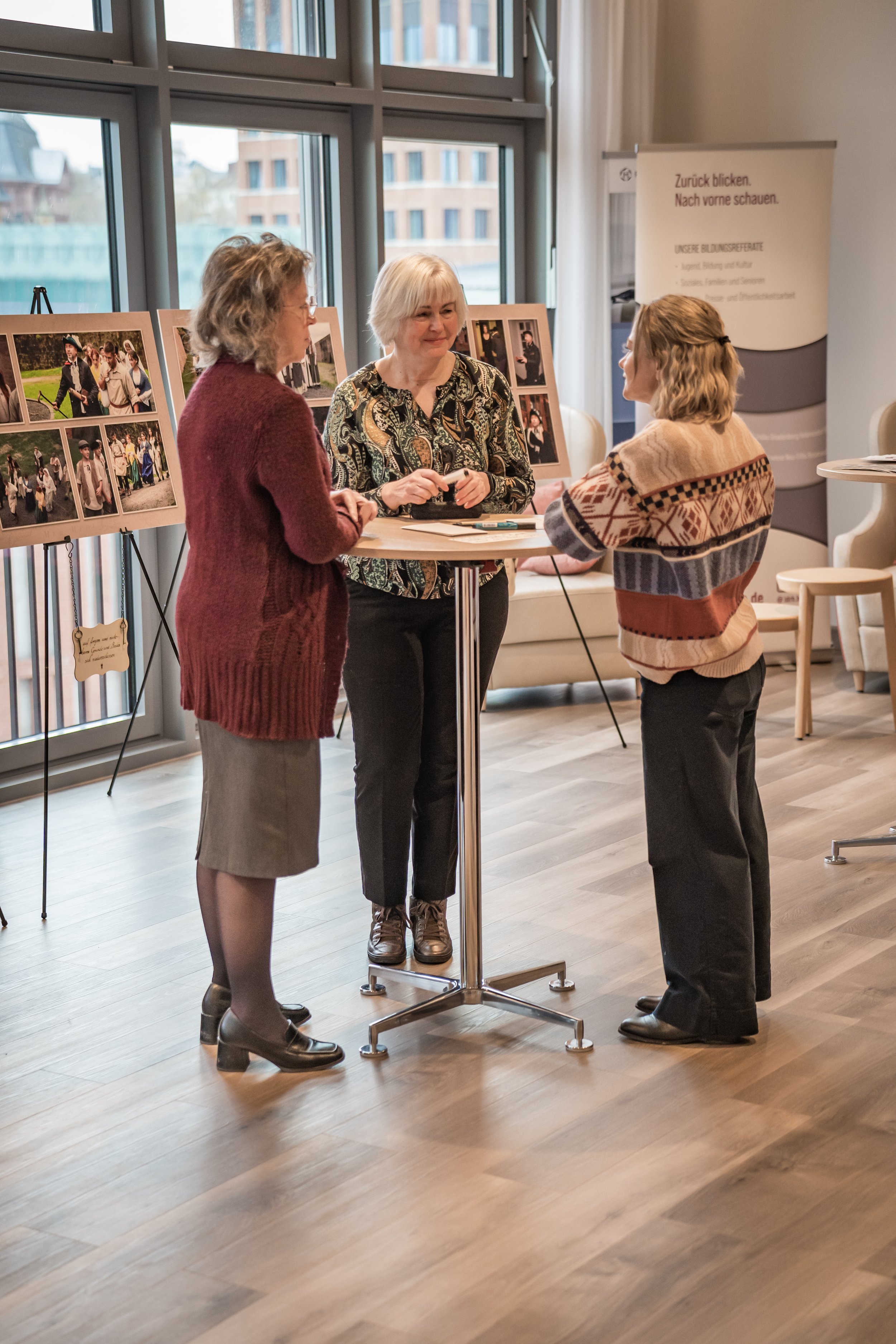 Drei Frauen unterhalten sich an einem Stehtisch in einem hell verständigen Raum mit großen Fenstern. Hinter ihnen sind Fotopräsentationen auf Ständern und ein Banner.