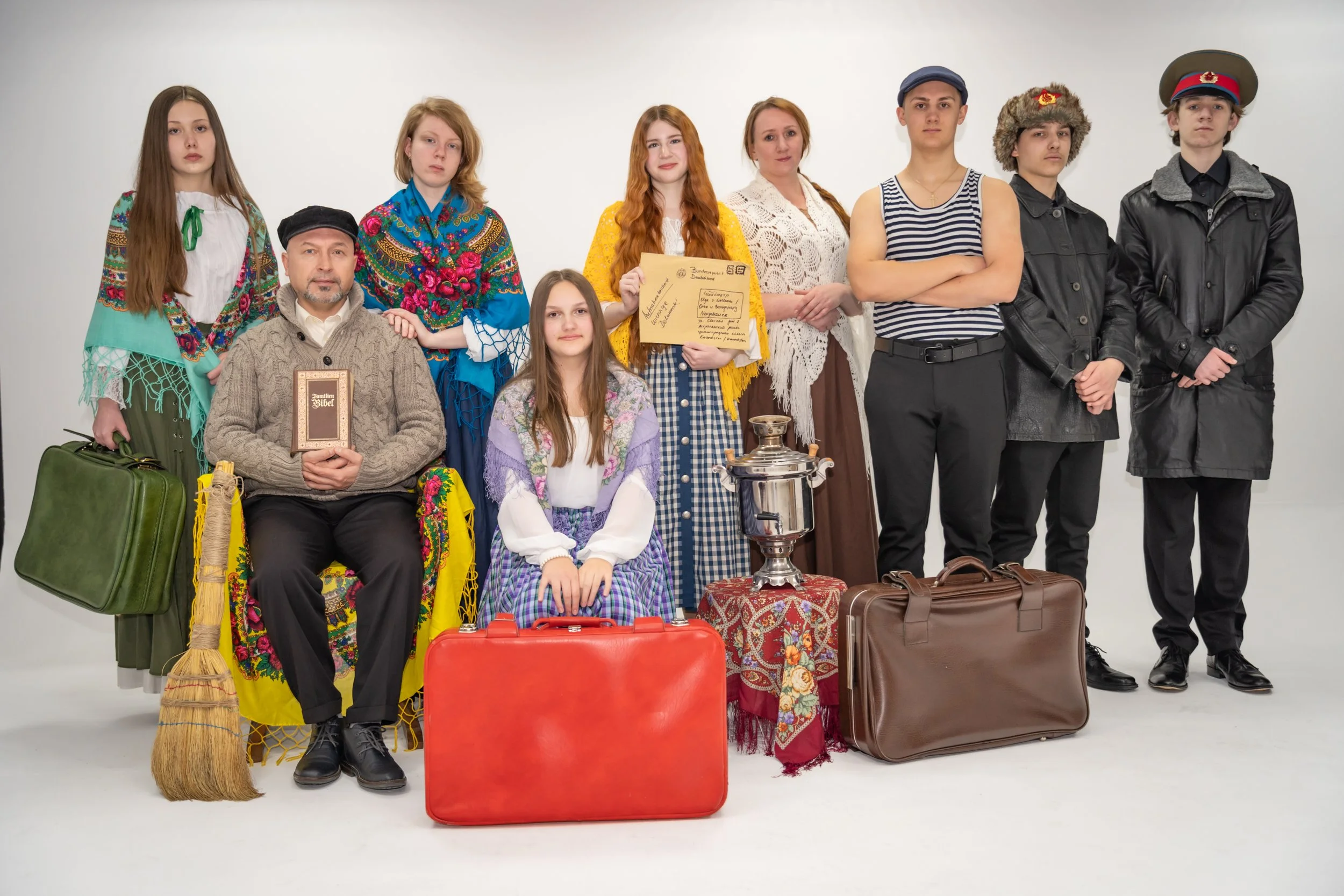 Gruppe von jungen Menschen und Erwachsenen in traditioneller Kleidung und Kostümen, vor einem weißen Hintergrund, mit Koffern und Requisiten, die auf eine Theater- oder Filmszene hindeuten.
