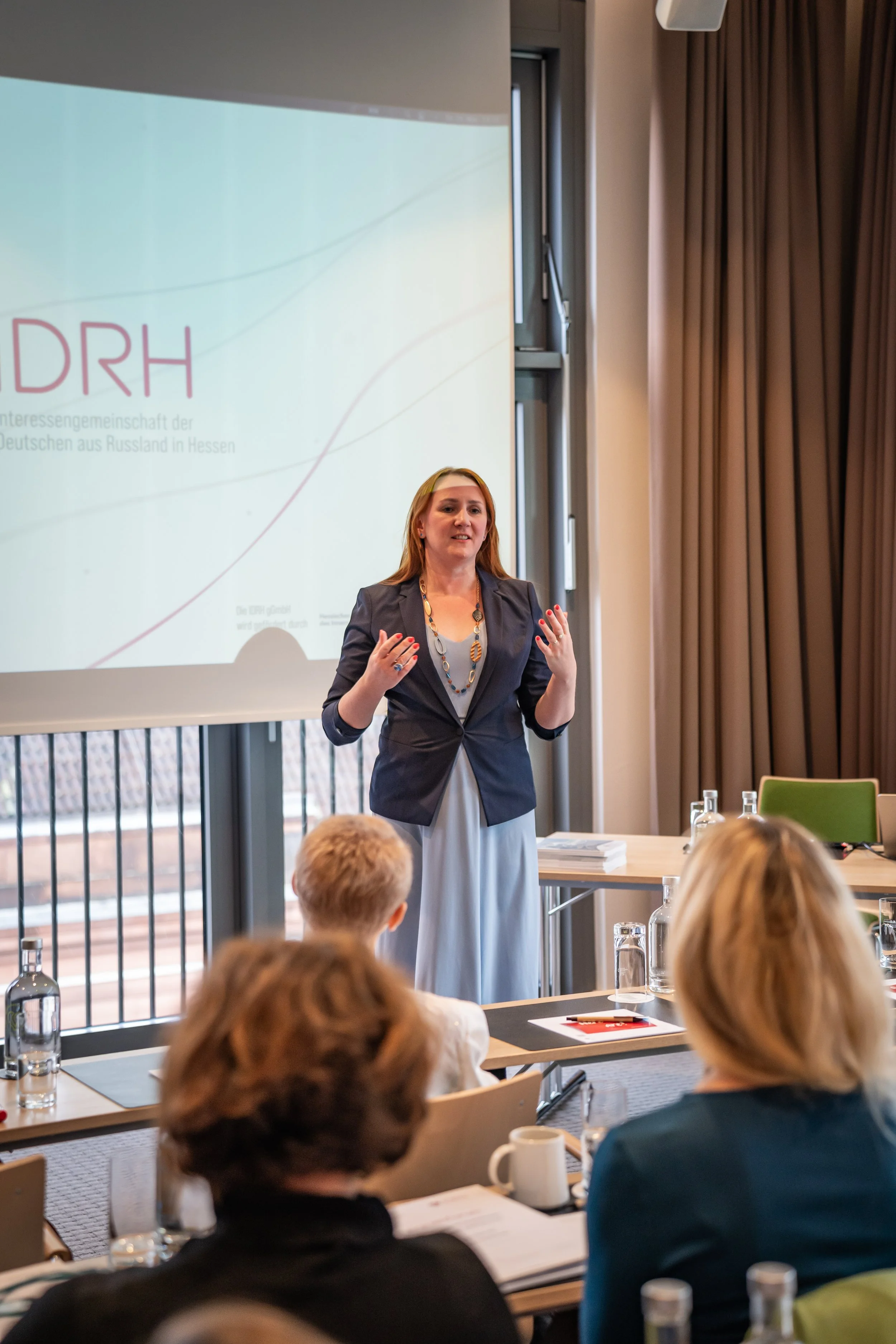 Eine Frau hält eine Präsentation vor einer Gruppe in einem Konferenzraum, mit einem Projektor im Hintergrund, der das Logo der Interessen Gemeinschaft der Deutschen aus Russland in Hessen zeigt.