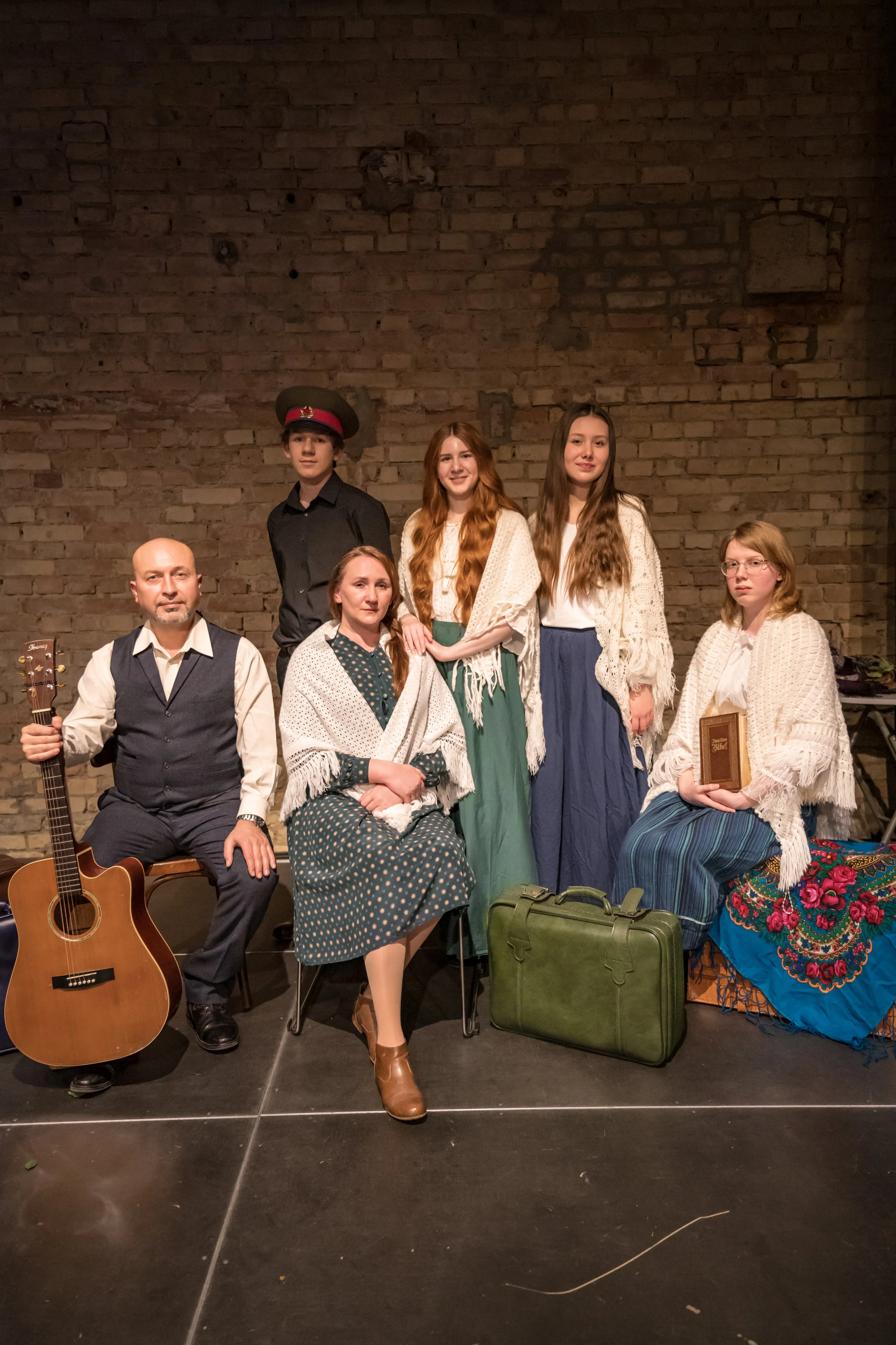 Gruppe von Menschen in Vintage-Kleidung, auf einer Bühne vor einer Backsteinmauer, mit einem Gitarrenkoffer und einem Aktenkoffer. Vorsitzende sitzt mit Papier und Handtasche, andere stehen hinten, eine hält ein Buch.
