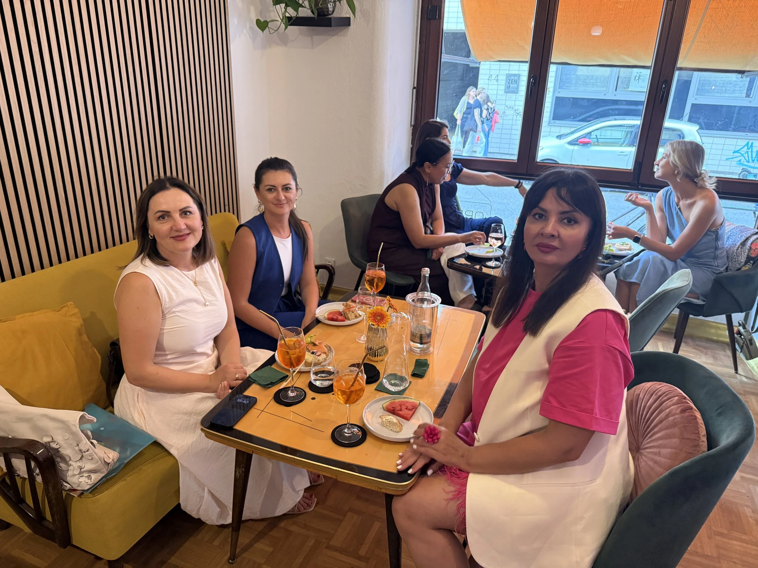 Vier Frauen sitzen an einem Holztisch mit Snacks und Getränken in einem modernen Café, einige sitzen auf einem gelben Sofa, andere auf Stühlen, im Hintergrund sitzen vier weitere Frauen bei einem Abendessen am Fenster.