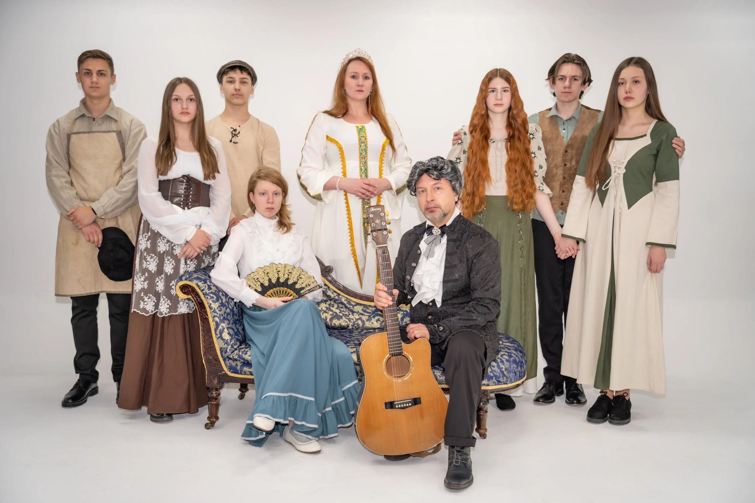 Eine Gruppe von Menschen in historischen Kostümen, mit einem Mann, der auf einem Stuhl mit Gitarre sitzt, und einer Frau, die auf einem Sofa mit Fächer sitzt, vor einem weißen Hintergrund.