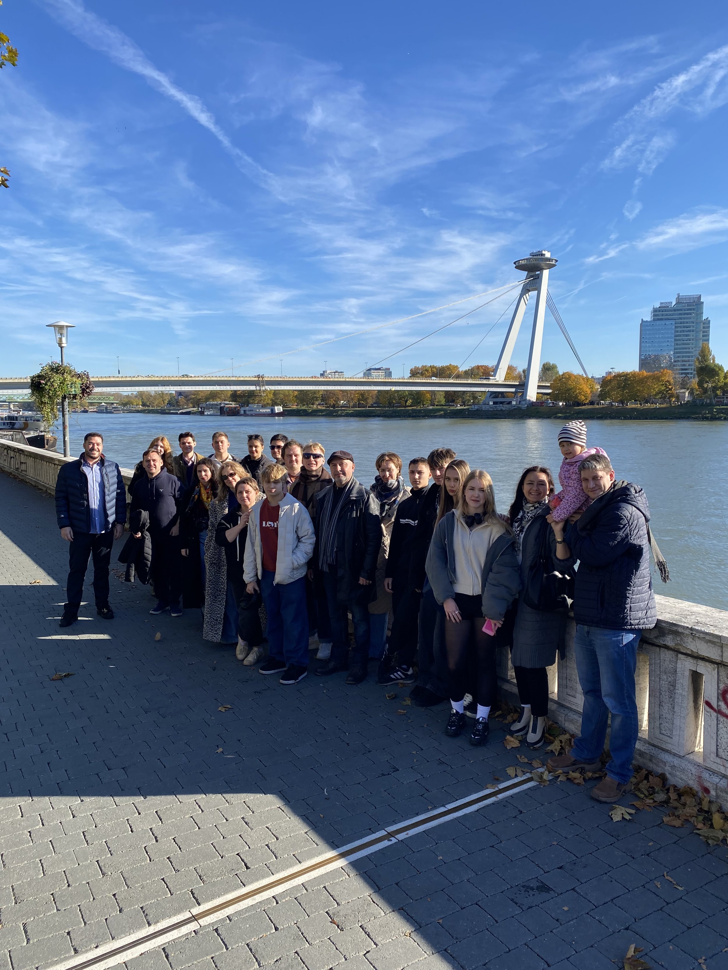 Eine Gruppe von Menschen steht an einem Flussufer mit einer Brücke und einem Fernsehturm im Hintergrund bei schönem Wetter.