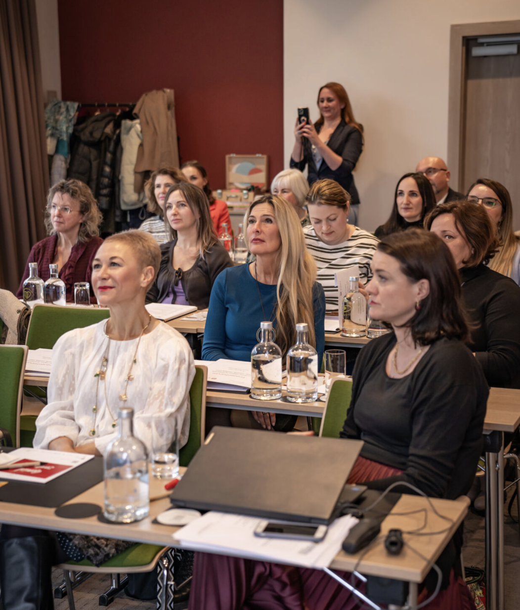 Mehrere Frauen sitzen bei einem Vortrag, eine Frau filmt die Veranstaltung mit dem Smartphone, im Hintergrund hängen Jacken an einer Garderobe.