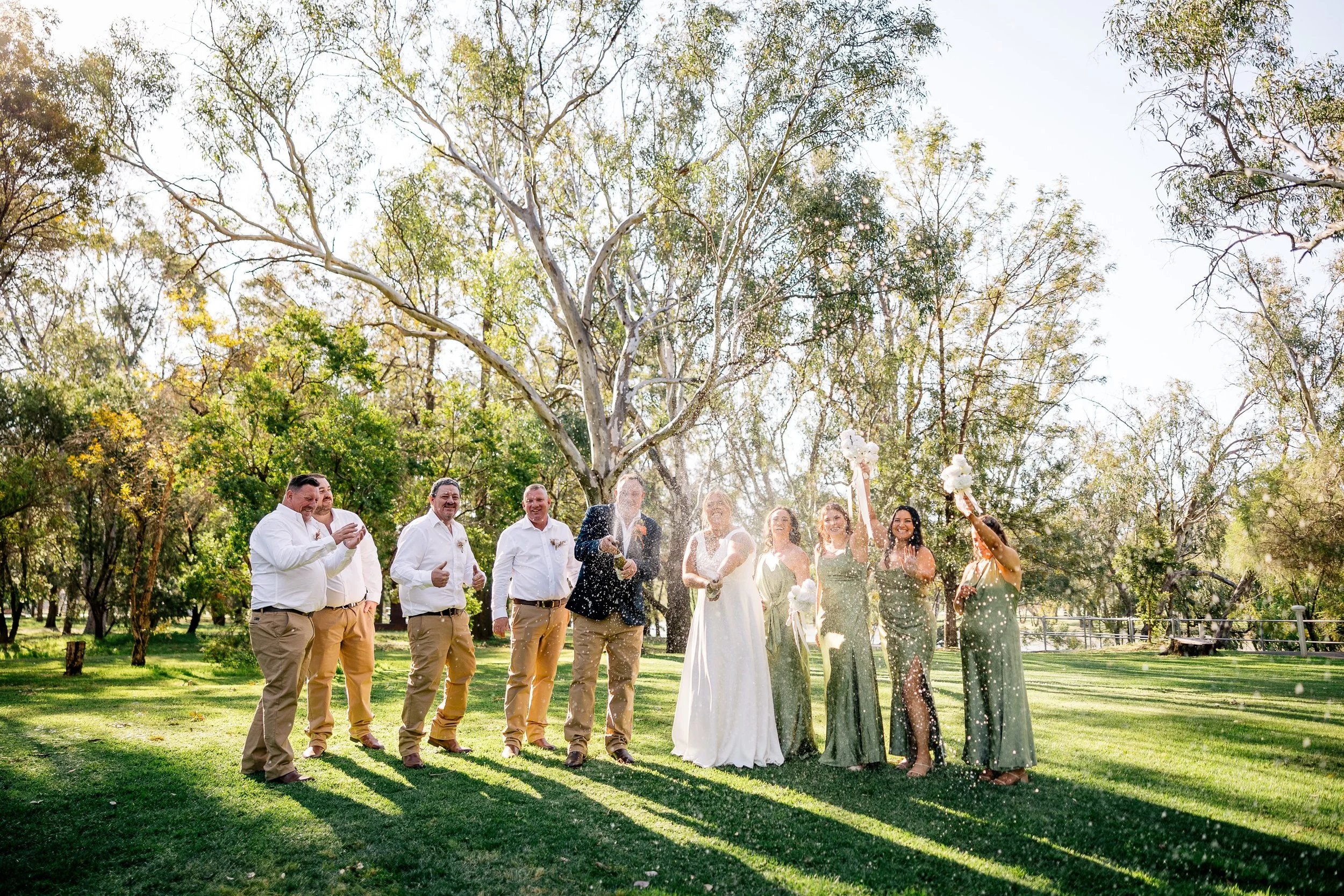 Group of people celebrating outdoors at a wedding, with some holding bouquets, dressed in formal attire, and making joyful gestures on grassy steps with trees in the background.