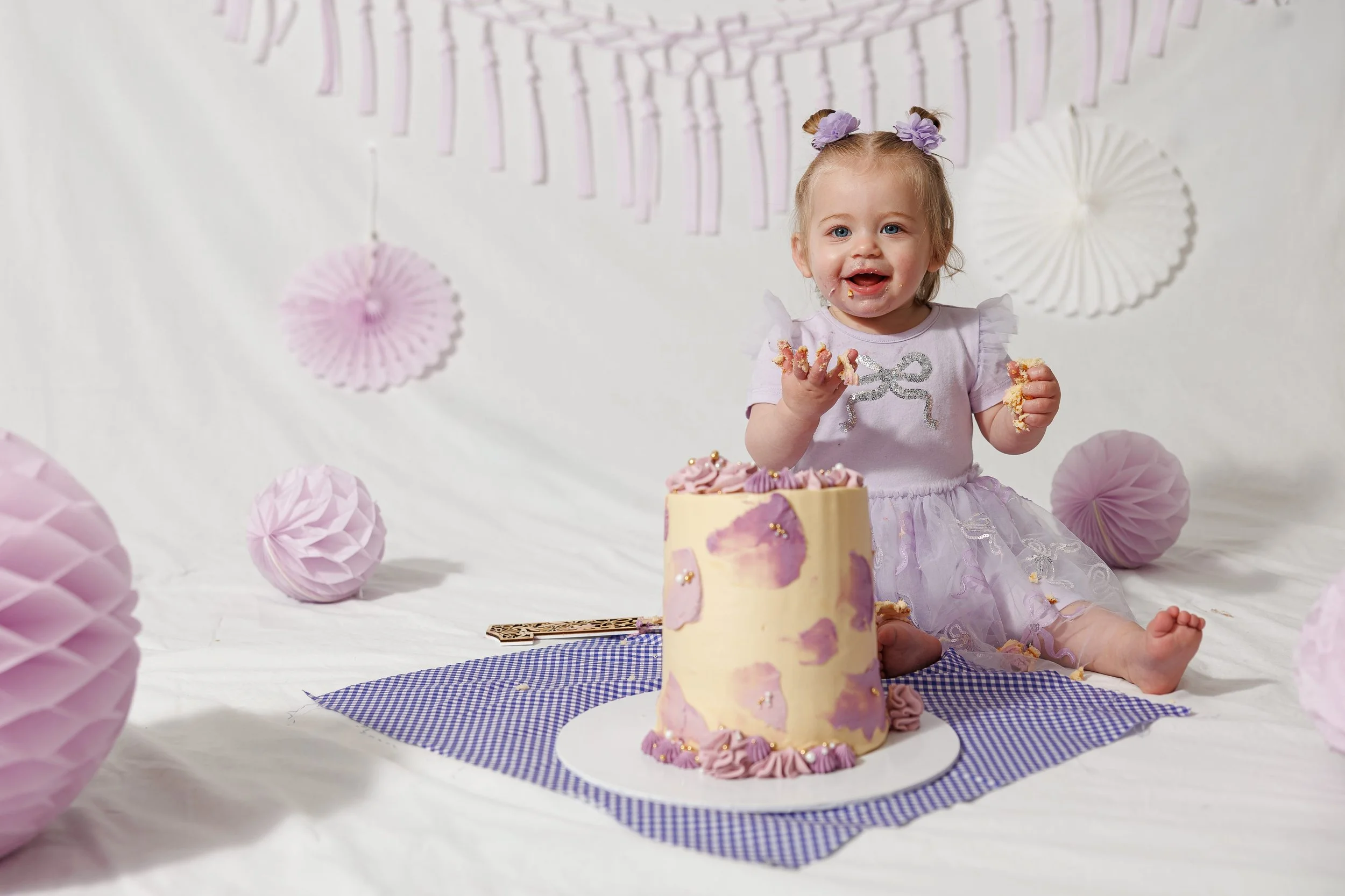 A smiling toddler girl in a pink tulle dress is sitting next to a partially eaten pink and white layered cake with pink decorations. She has cake and frosting on her face, hands, and clothes, and appears to be happily enjoying the moment. Pink paper lanterns are hanging in the background.