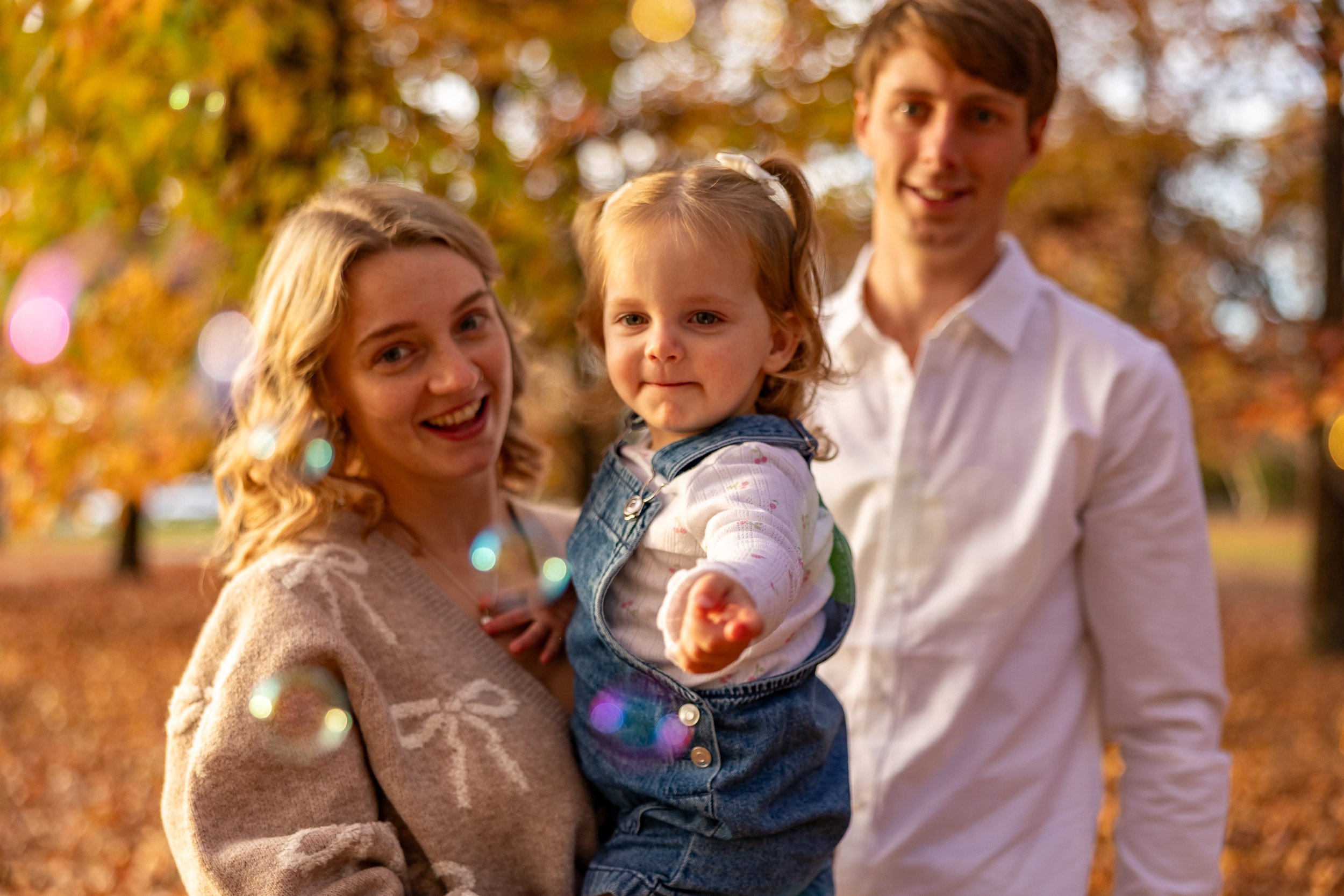 A happy family of three members, a mother, a father, and their young daughter, outdoors in an autumn park. The mother, with blonde wavy hair, is holding the daughter, who is pointing towards the camera and smiling. The father, with light brown hair, is standing behind them. The background is filled with fall foliage and warm sunlight.