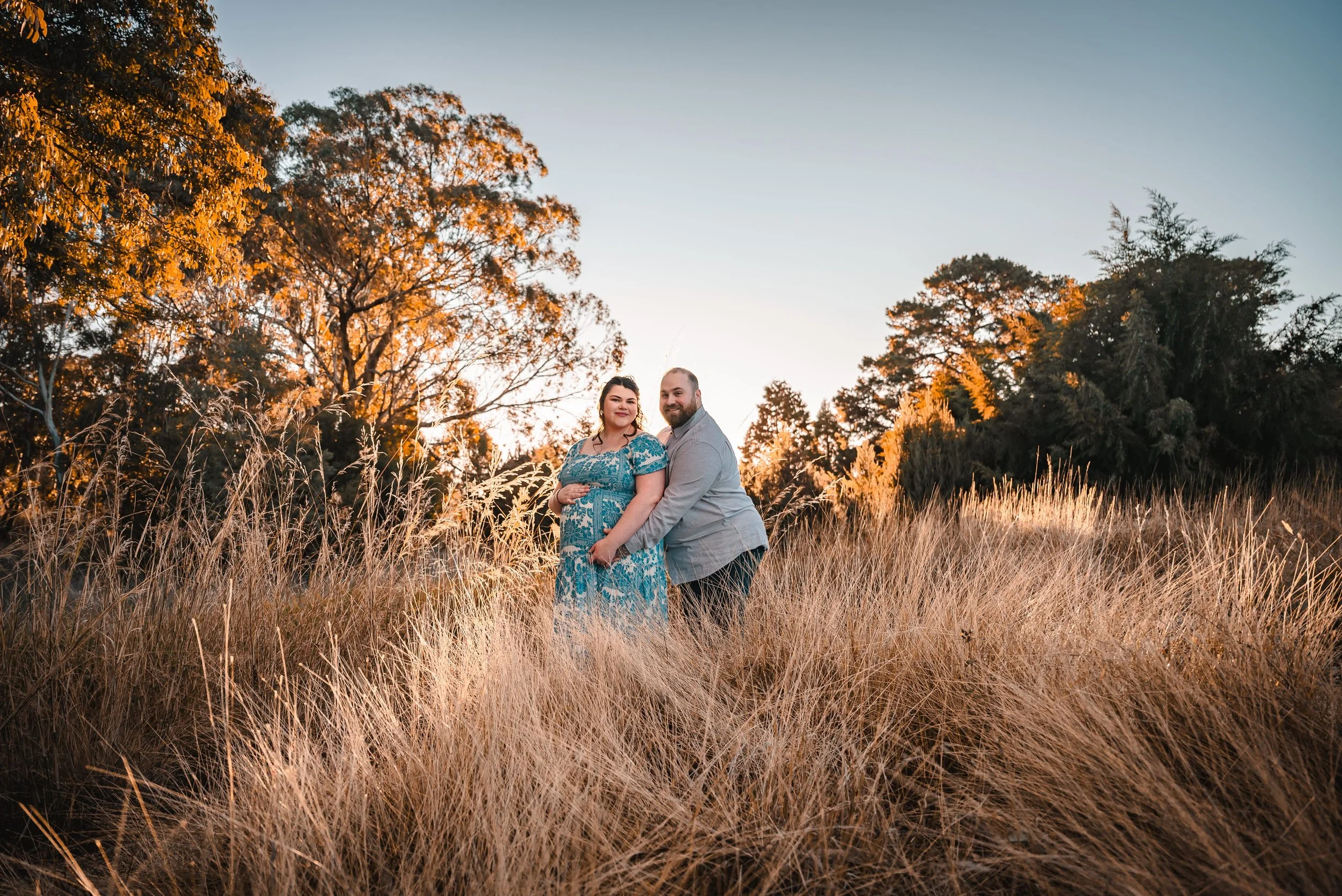 A couple standing in tall, dry grass during sunset in a natural outdoor setting, with trees in the background.