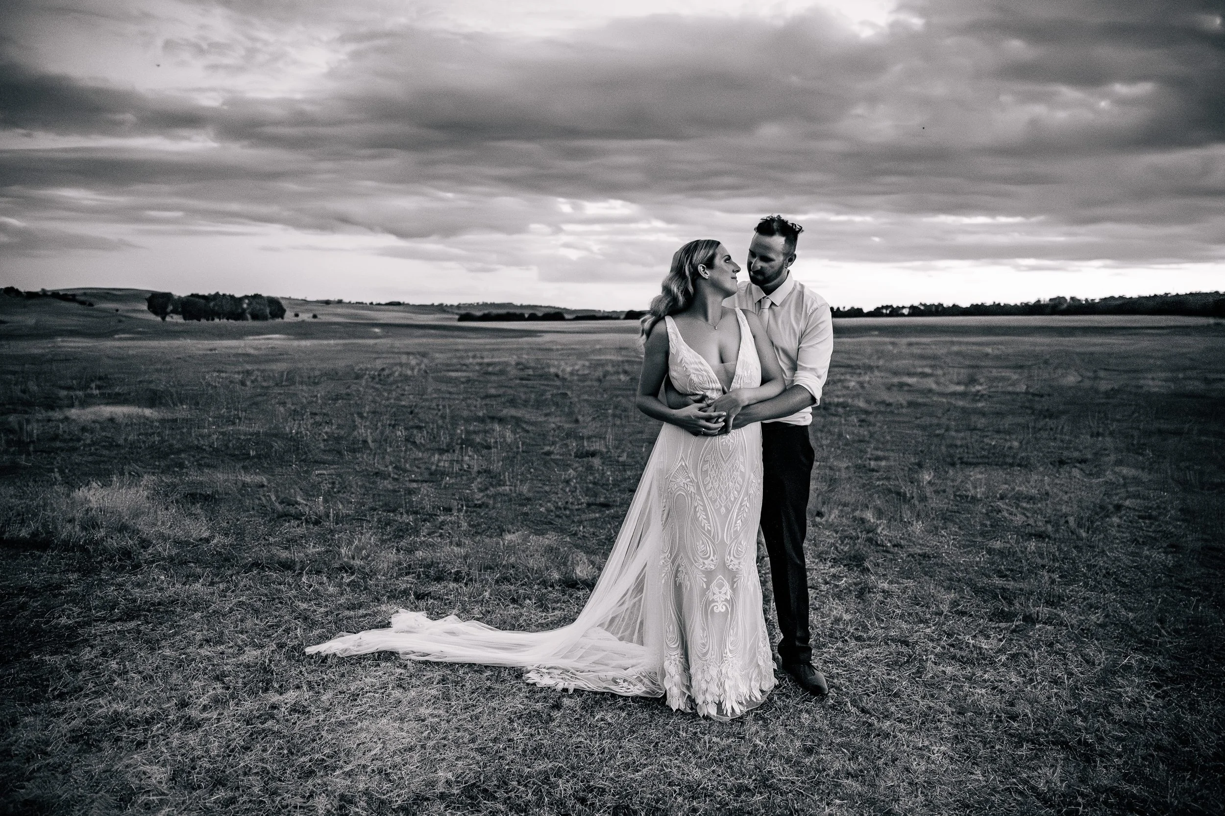A large wedding celebration outdoors with a bridge and water in the background, featuring a bride and groom surrounded by guests on a grassy field.
