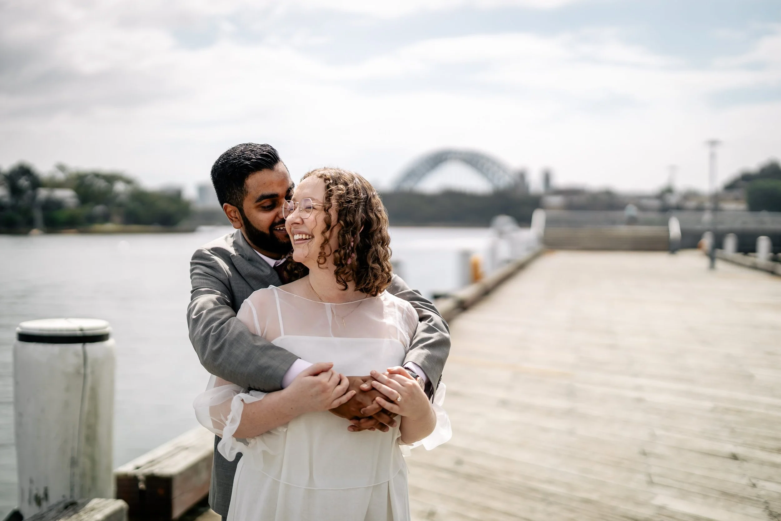 A couple dressed in wedding attire walking hand in hand across a wide open field at dusk, with a cloudy sky overhead.