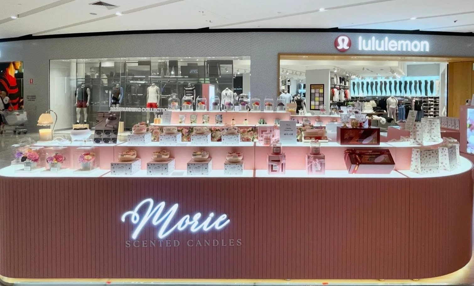 A store kiosk in a shopping mall selling scented candles with a pink display counter and decorative flowers, located in front of an American Eagle and lululemon store.