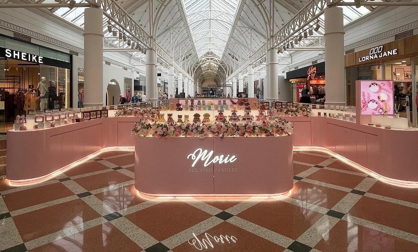 Inside a shopping mall with a pink display kiosk for Mui scented candles, decorated with flowers. Surrounding shops include Sheike, Lorna Jane, and others, with shoppers walking through the bright, glass-roofed corridor.