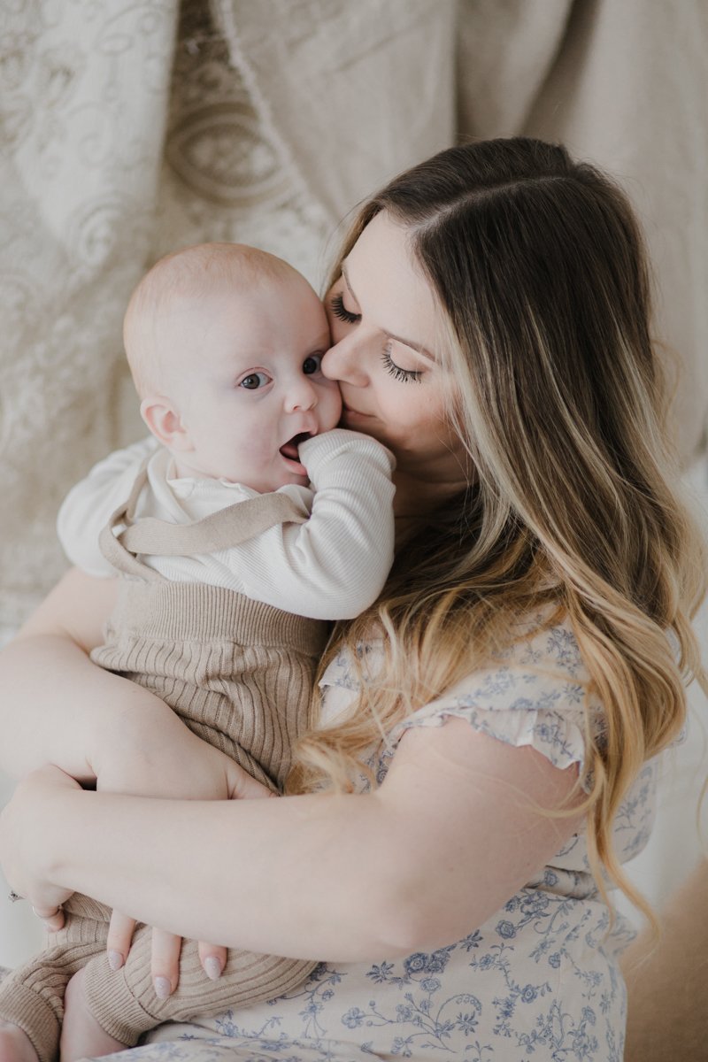 A woman cuddling a baby, kissing the baby's cheek, with a neutral background.