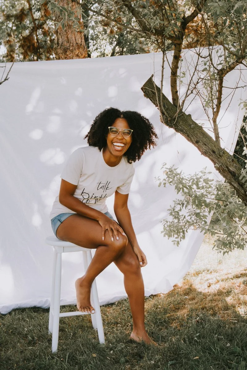 A woman with curly hair and glasses sitting on a white stool outdoors. She is smiling, wearing a white t-shirt and denim shorts, and sitting with one leg crossed over the other. There is a tree and a white cloth backdrop behind her, with sunlight filtering through the leaves.