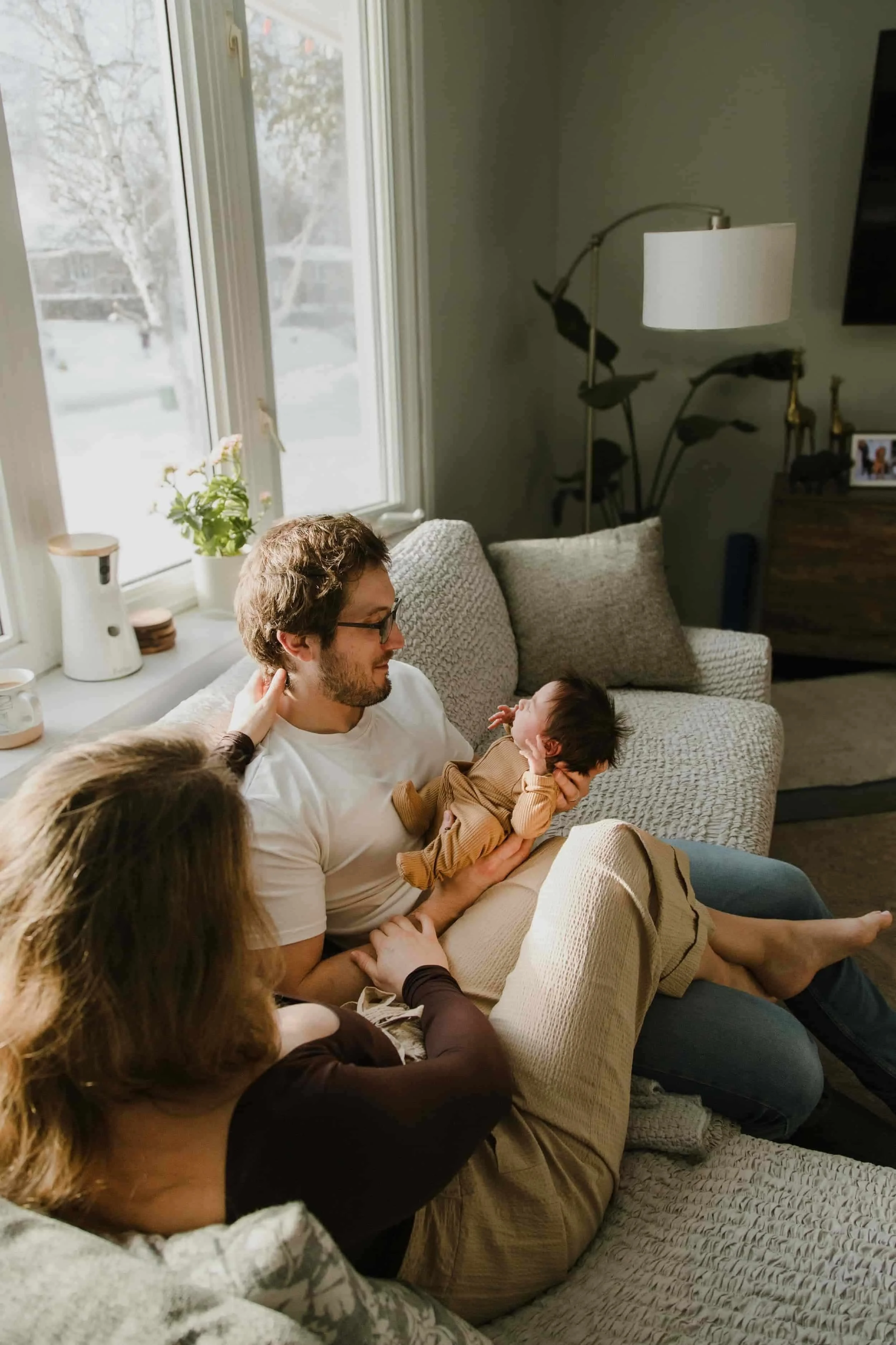 Family sitting on a couch in a living room, with a man holding a baby, and a woman touching the man's face, near a window with snowy scenery outside.