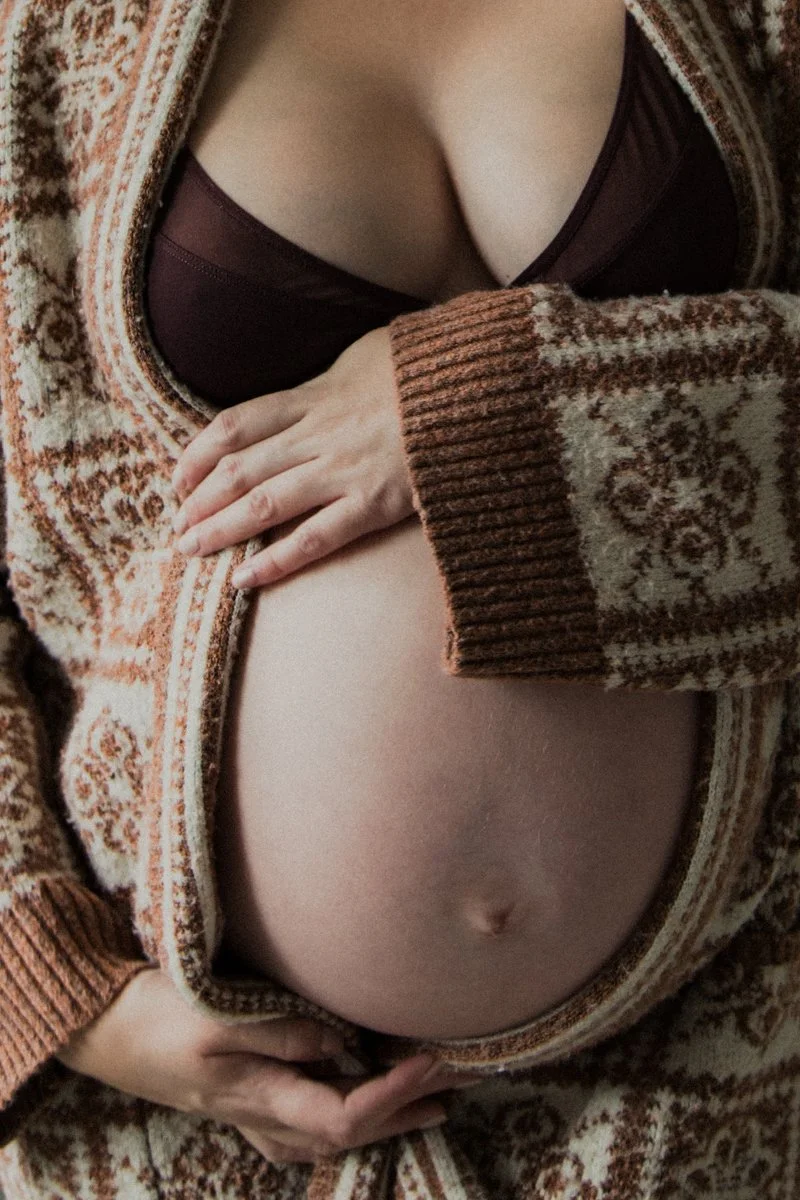 Close-up of a person's bare midsection, with one hand resting on their stomach and the other hand holding the edge of a patterned, brown, and beige cardigan.