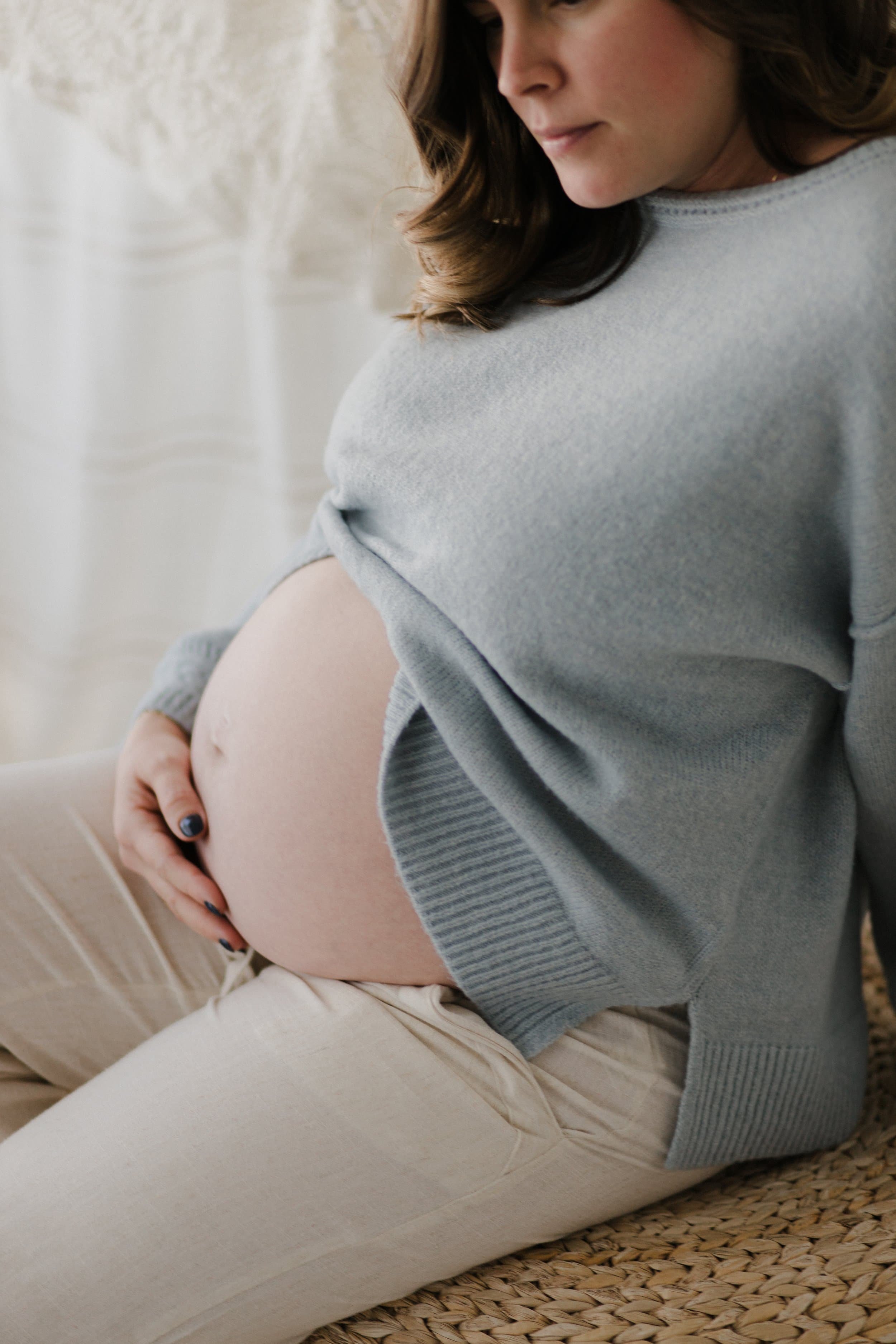 A pregnant woman sitting on a woven mat, gently holding her belly, in studio by Oakville family photographer, Stephanie Collin.
