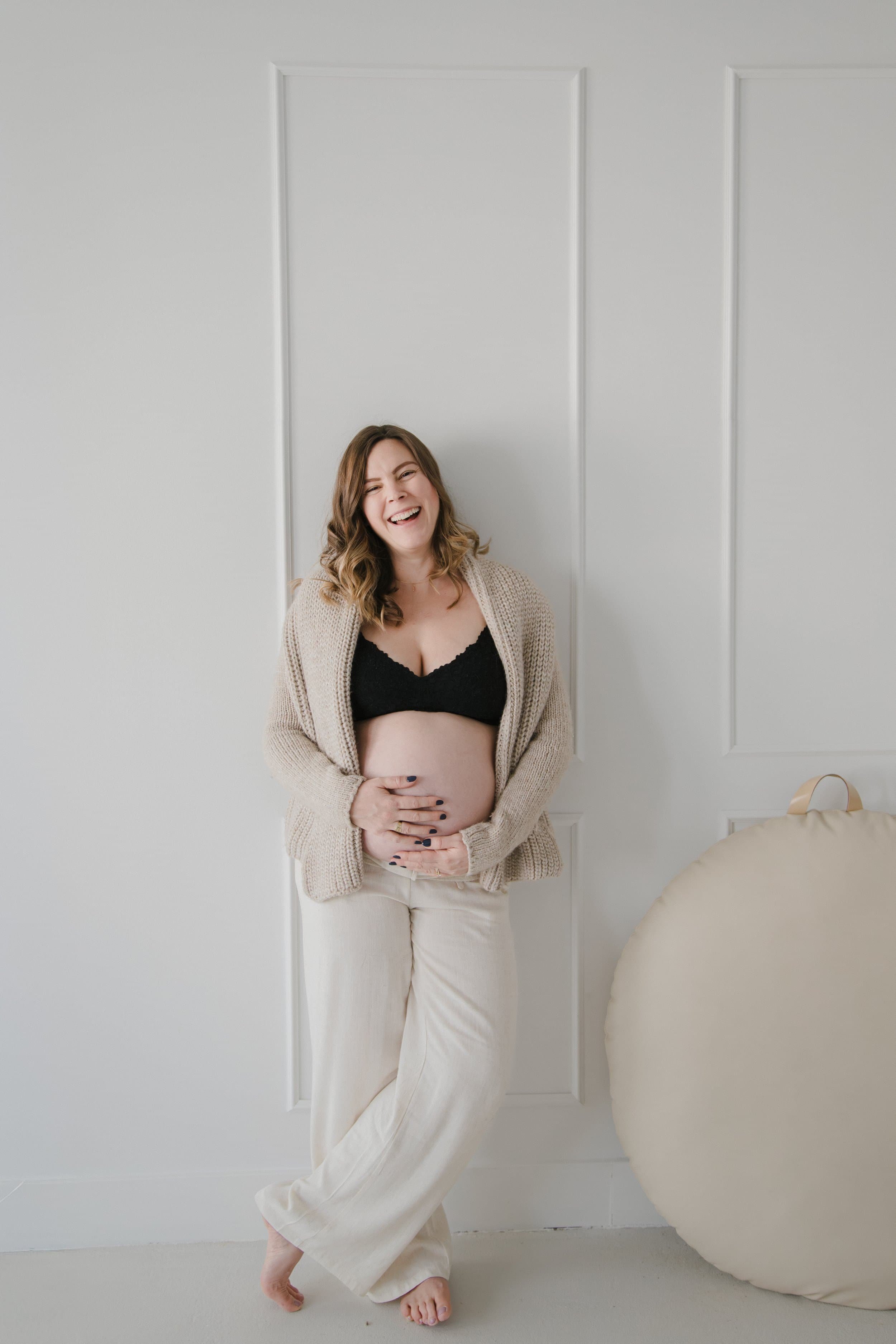 Smiling pregnant woman with shoulder-length brown hair, wearing a black lace bra, beige cardigan, and cream-colored pants, standing barefoot against a white wall, holding her baby bump. Oakville Studio Session