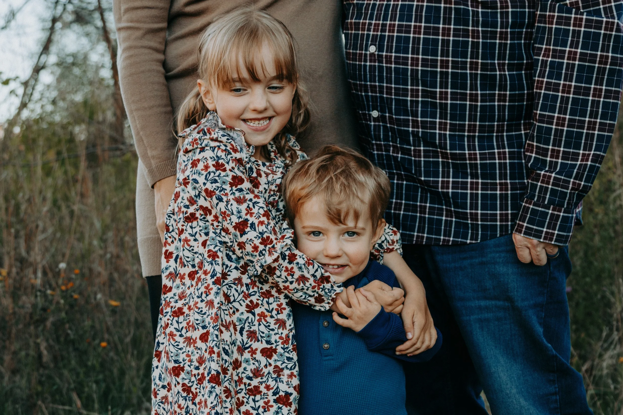 A family of four outdoors, with two children hugging, the girl smiling and the boy looking at the camera, with partial views of two adults behind them.