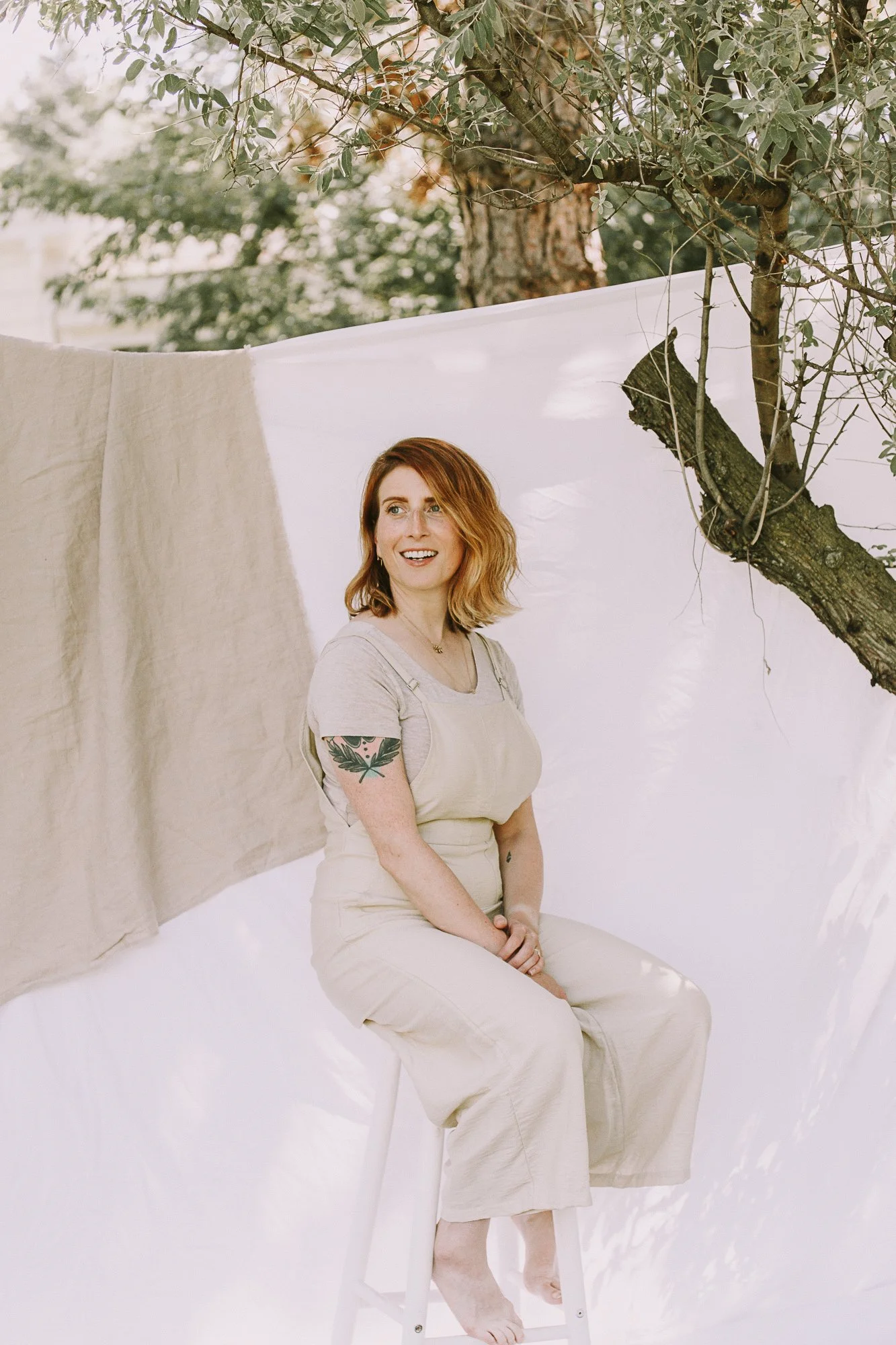 A woman with red hair, tattoos, and a beige overalls sitting barefoot on a stool outdoors, with fabric and trees behind her.