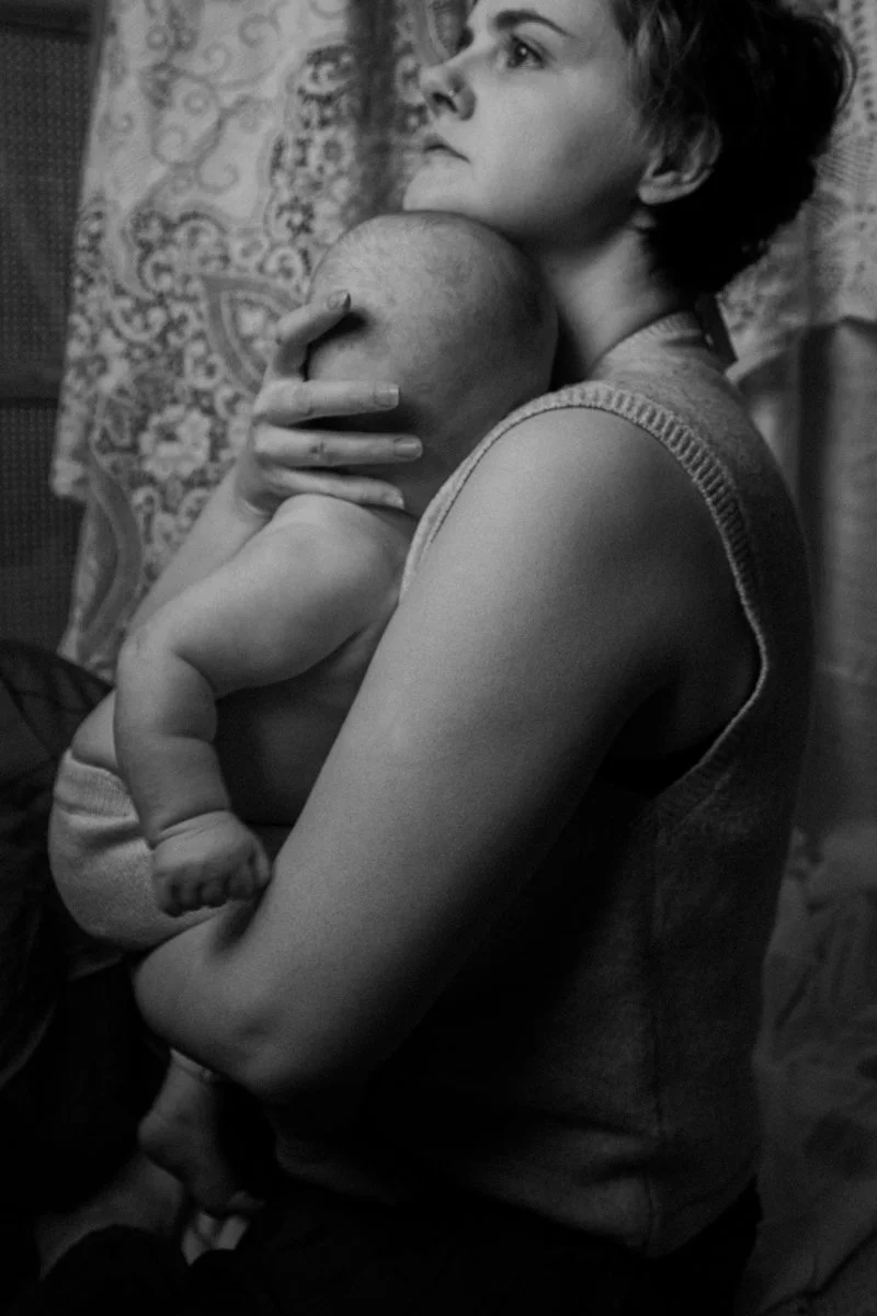A woman holding a baby close to her chest, looking thoughtfully to the side in a domestic setting.