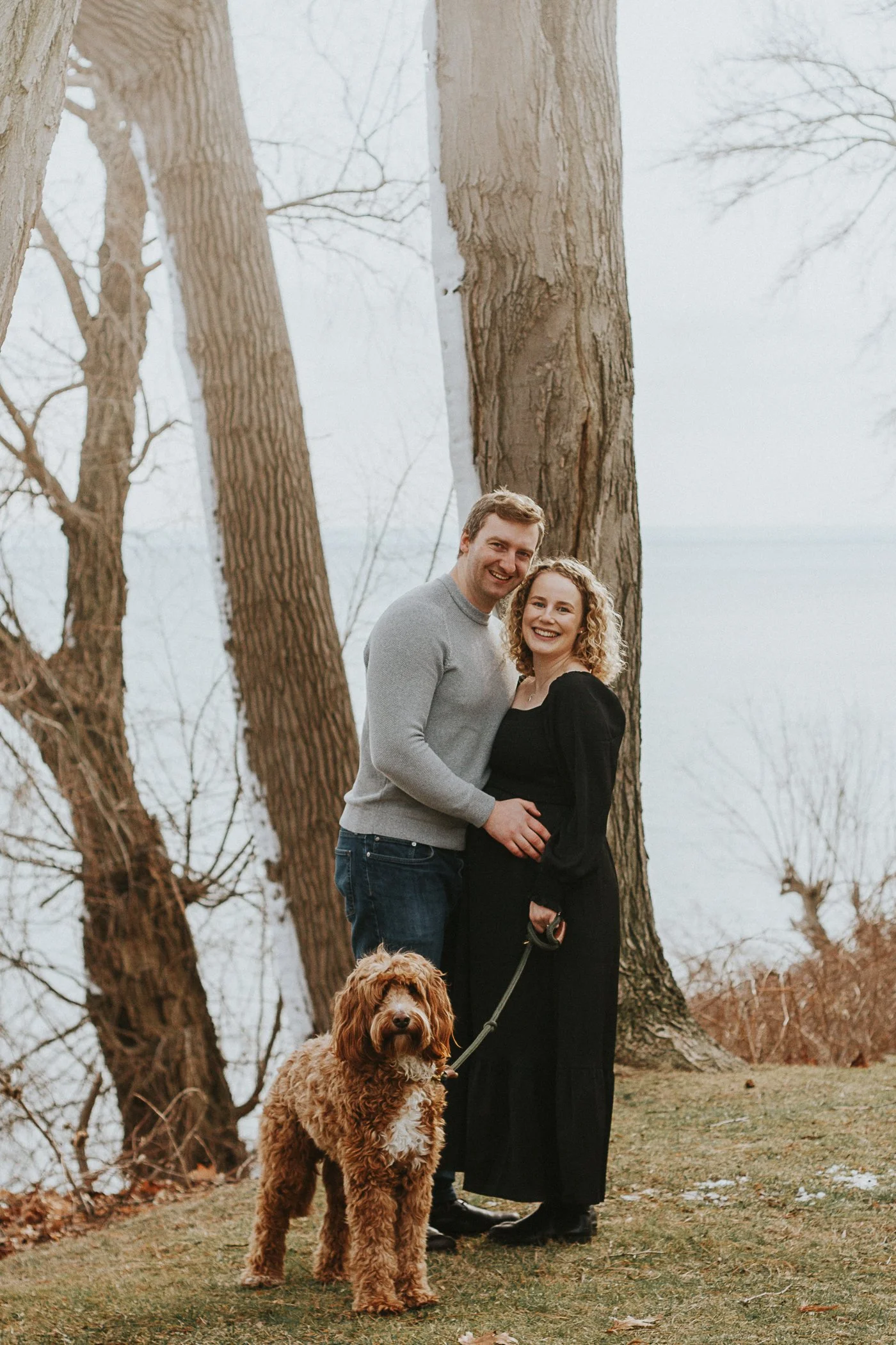 A smiling couple standing outdoors near a large tree, with the pregnant woman holding a leash attached to a brown curly-haired dog.
