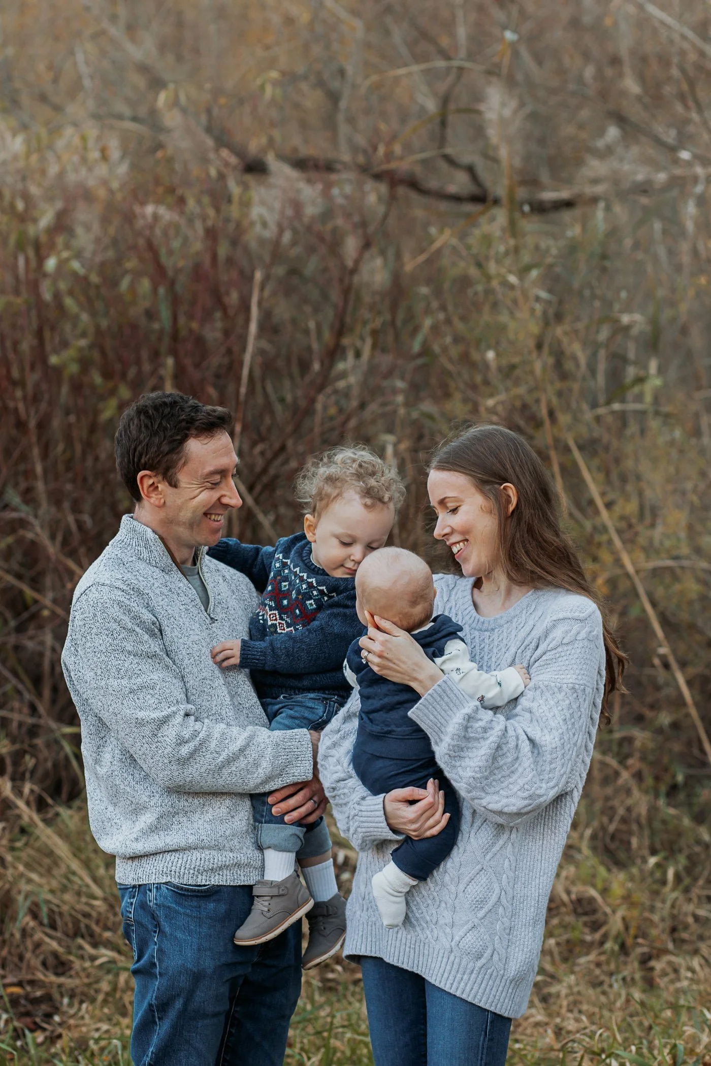 A smiling family of four hugs outdoors; captured by an Oakville family photographer, they're surrounded by trees and greenery.