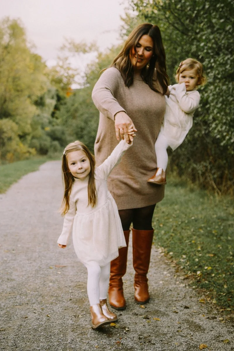 A woman walking outdoors on a path with two young girls, holding hands with the girl in front, and carrying the girl in back, surrounded by trees and greenery.