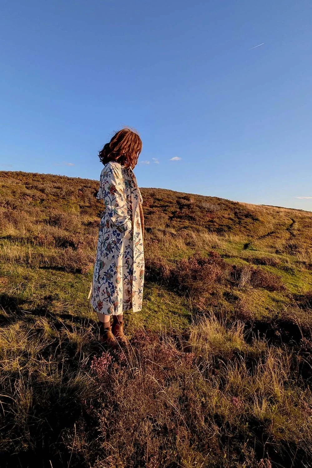 Woman wearing a floral print trench coat made from vintage fabrics, stood side on in the beautiful North Yorkshire Moors backdrop.