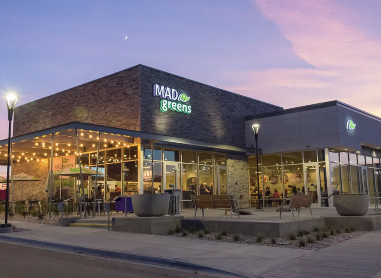 Exterior of a modern restaurant called MAD Greens during sunset with outdoor seating, string lights, and a lit sign, against a sky with a crescent moon.