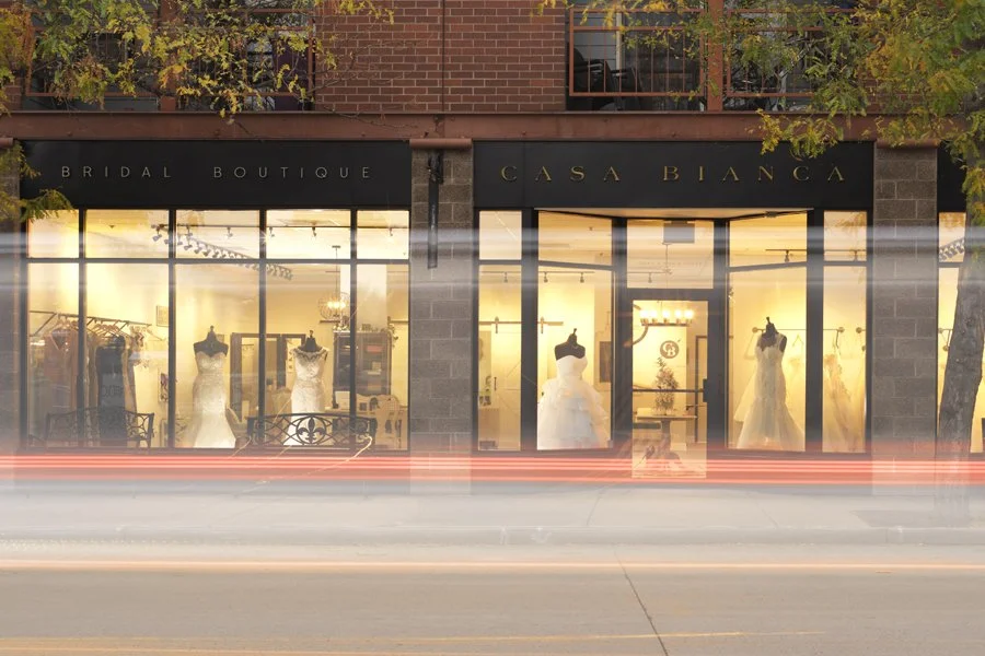 Storefront of Casa Bianca bridal boutique with wedding dresses on display inside, illuminated and visible through large glass windows, located on a city street with streaks of light from passing cars.