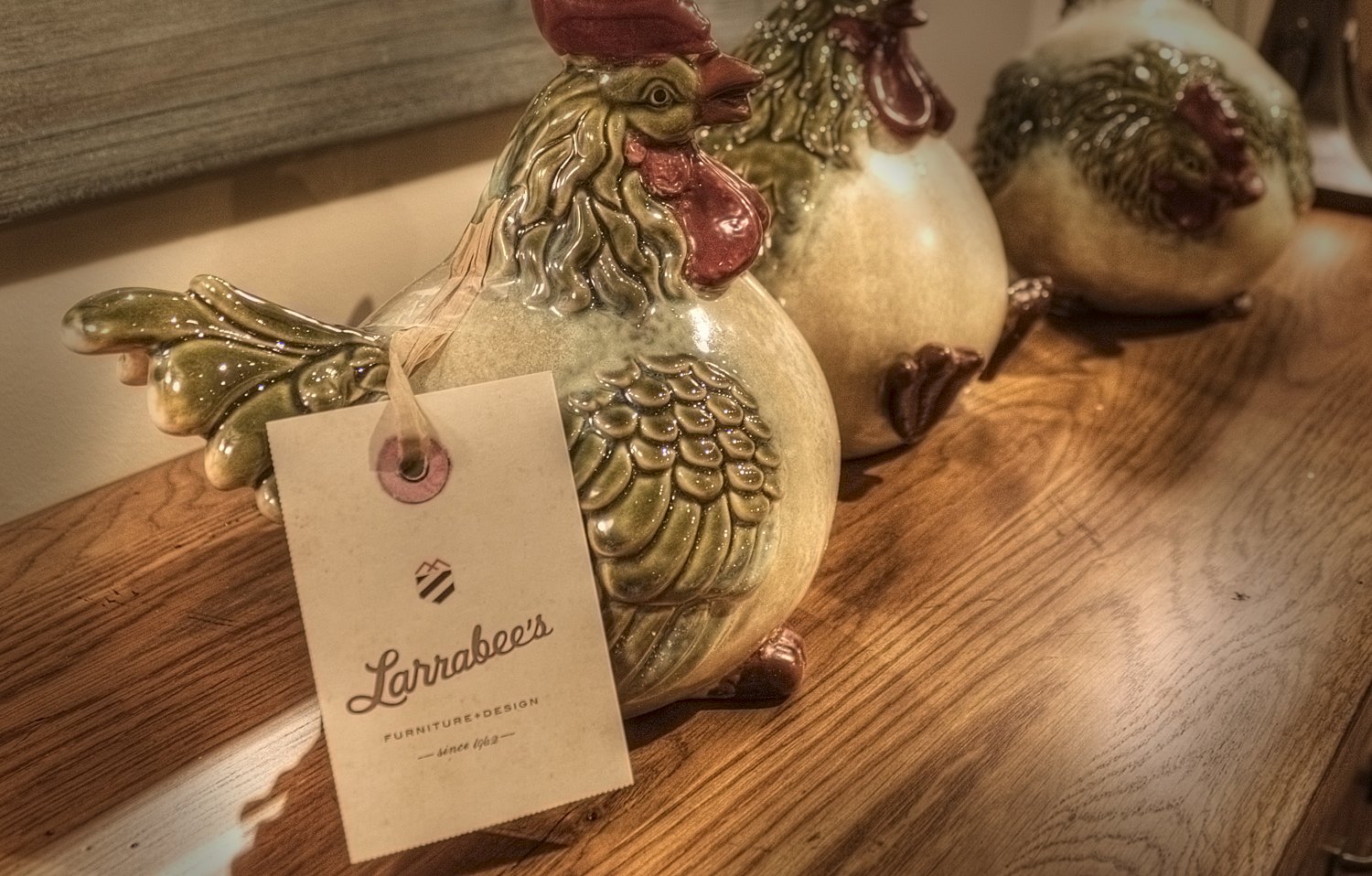Ceramic figurines of a rooster and hens on a wooden surface, with a tag reading "Larrabees Furniture + Design since 1962."