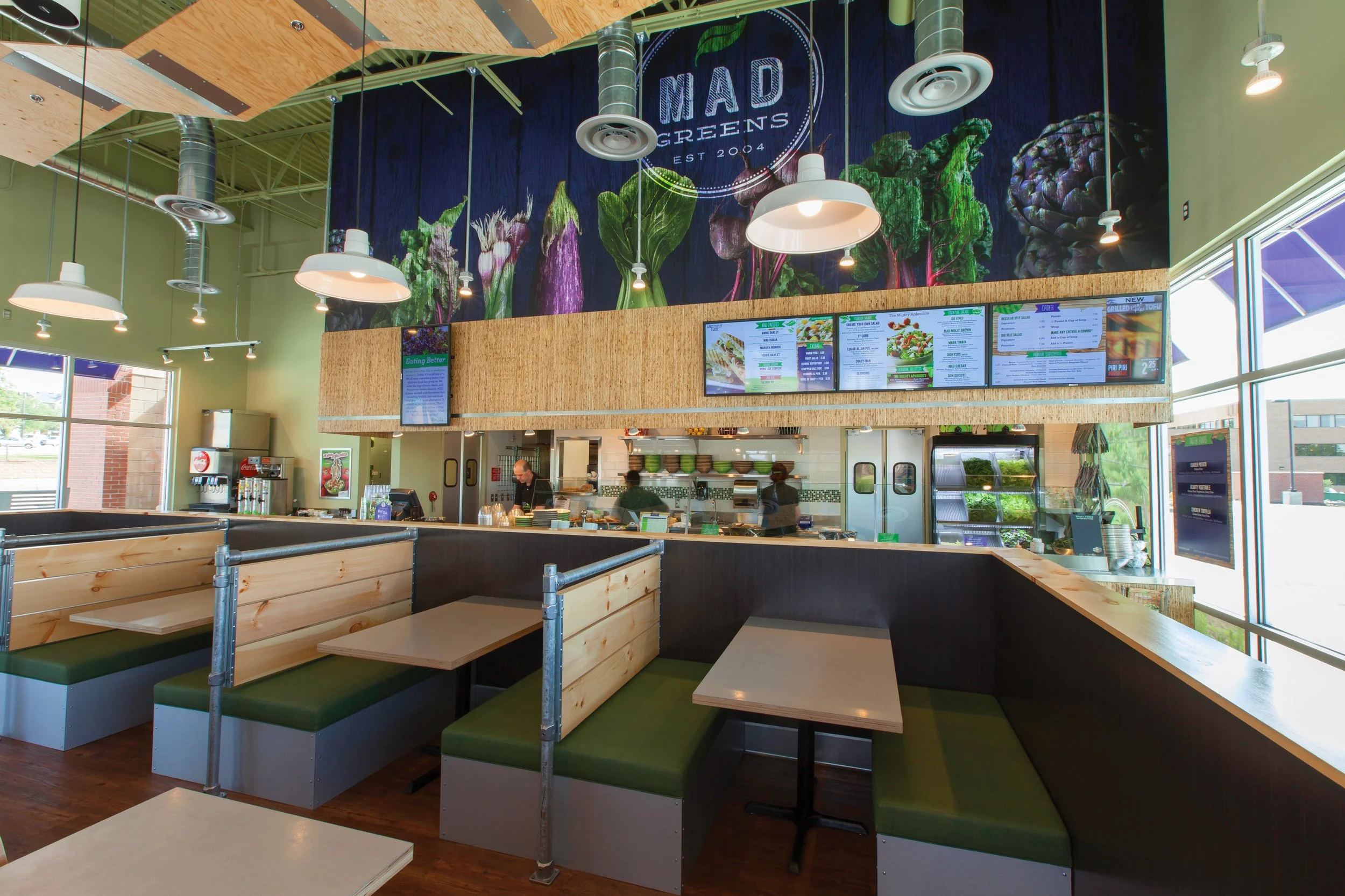 Interior of a healthy fast casual restaurant with green booths, digital menu boards, and a counter for orders, branded as MAD Greens, featuring a large wall graphic of vegetables.
