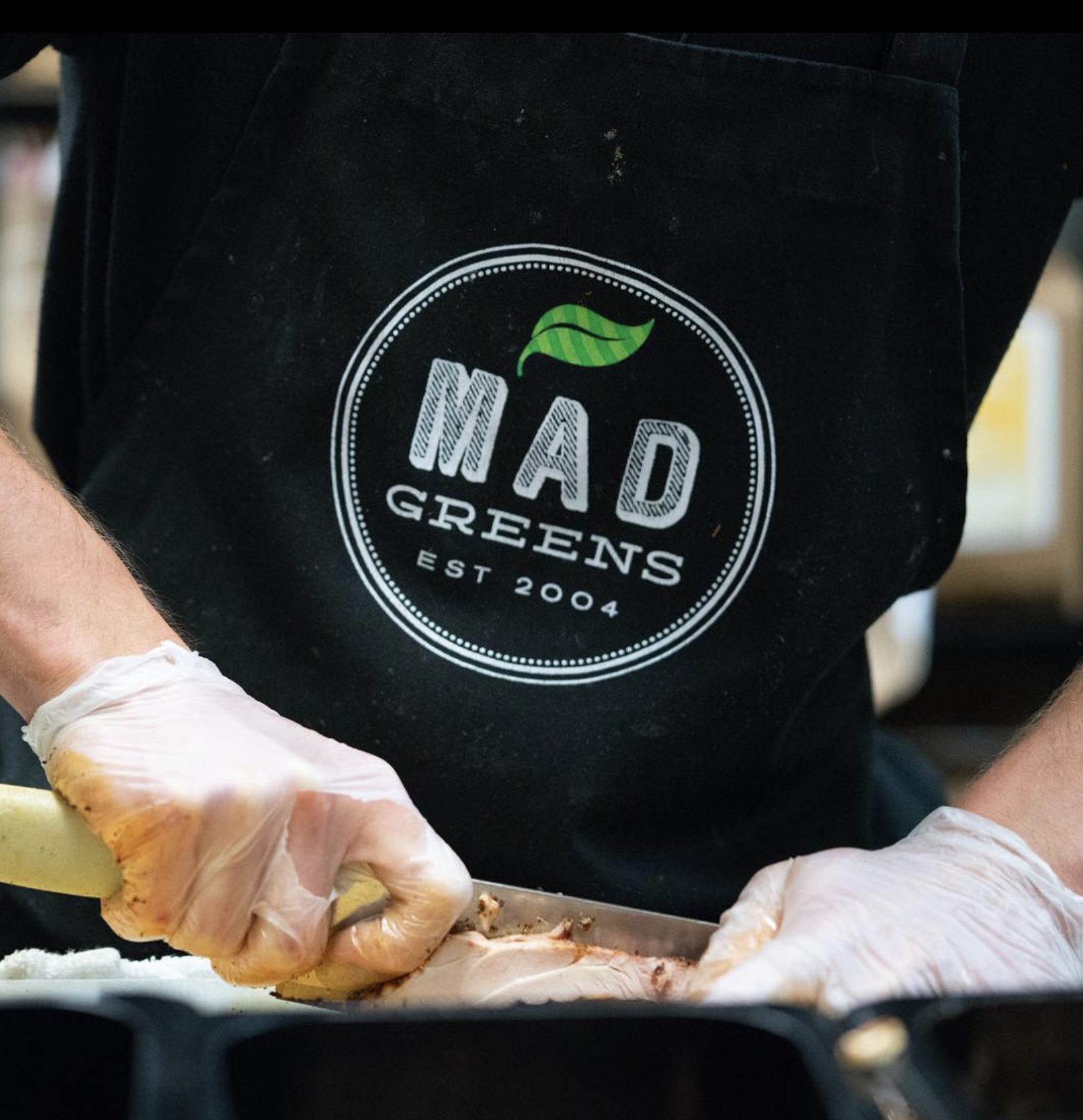 Person wearing a black apron with the logo 'I Mad Greens' and the establishment year 2004, preparing food with gloves on.