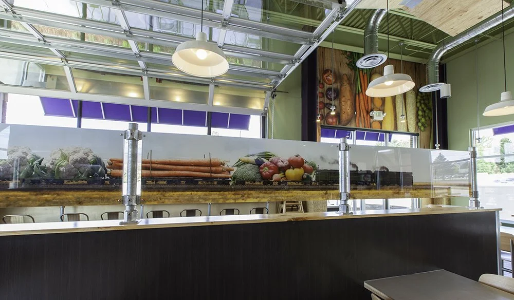 Display of fresh vegetables including carrots, cauliflower, broccoli, and peppers behind a glass barrier in a restaurant or cafe setting.