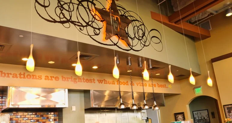Interior of a restaurant with hanging lights, a decorative star with swirling wire around it on the ceiling, and a quote on the wall that reads 'Celebrations are brightest when good food & vino are best'.