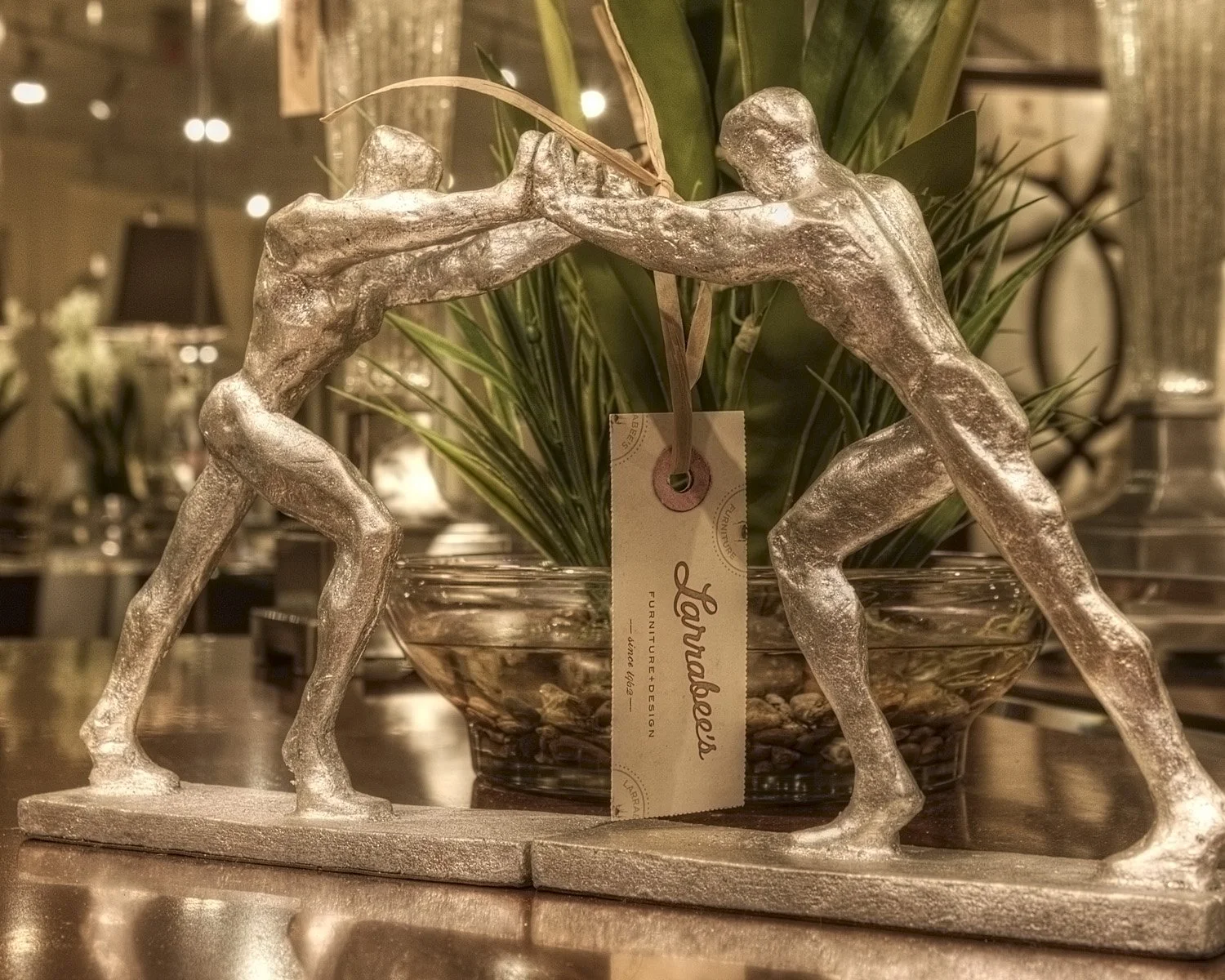Decorative silver sculpture of two wrestlers grappling, placed in front of a glass bowl with green plants, on a wooden surface.