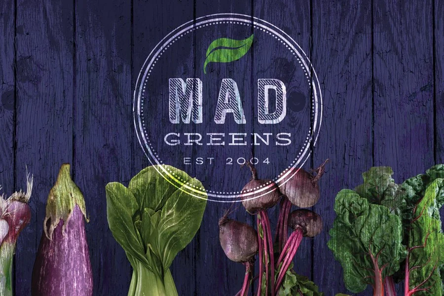 Logo for Mad Greens with a green leaf above the name, surrounded by fresh vegetables including purple eggplant, green lettuce, beets, and Swiss chard on a purple wooden background.