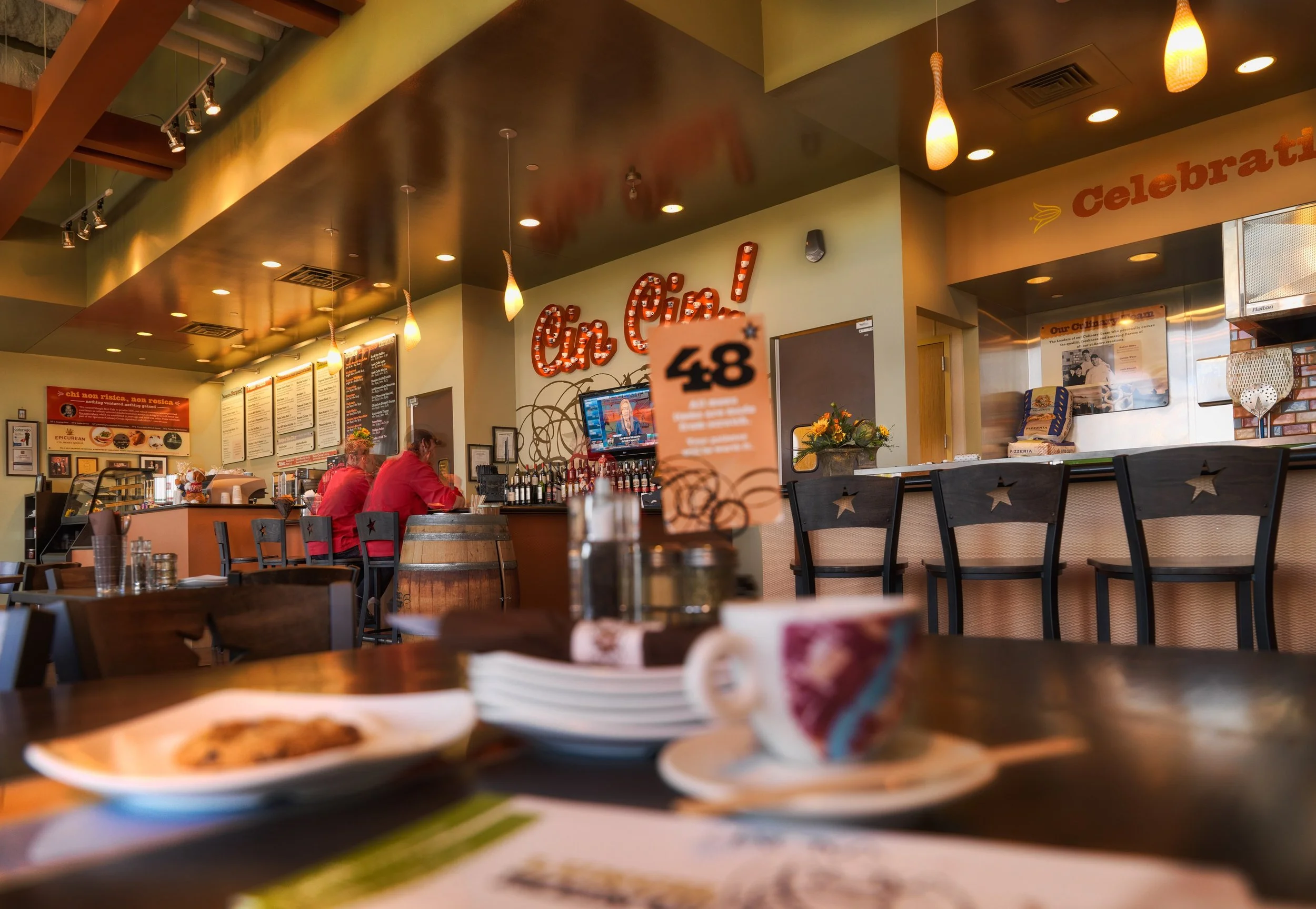 Interior of an Italian restaurant with a bar, bar stools, and a TV. Tables with plates of food and a coffee mug are in the foreground. Warm lighting and decorative signs are visible.