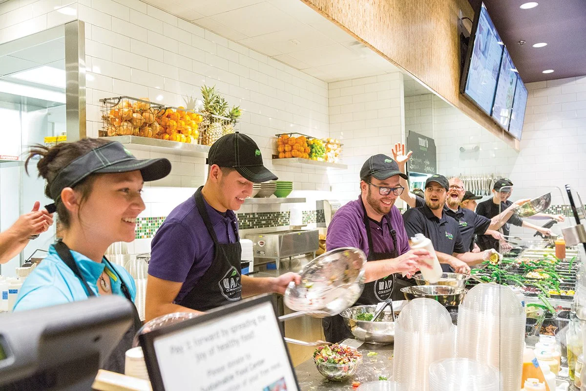 A group of workers in a restaurant kitchen preparing food and smiling, wearing aprons and hats.