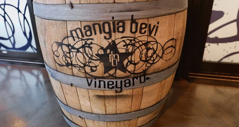 Wooden barrel with the logo 'Mango devi Vineyard' and a star, vine illustrations, located indoors with a wooden floor and decorative panels.