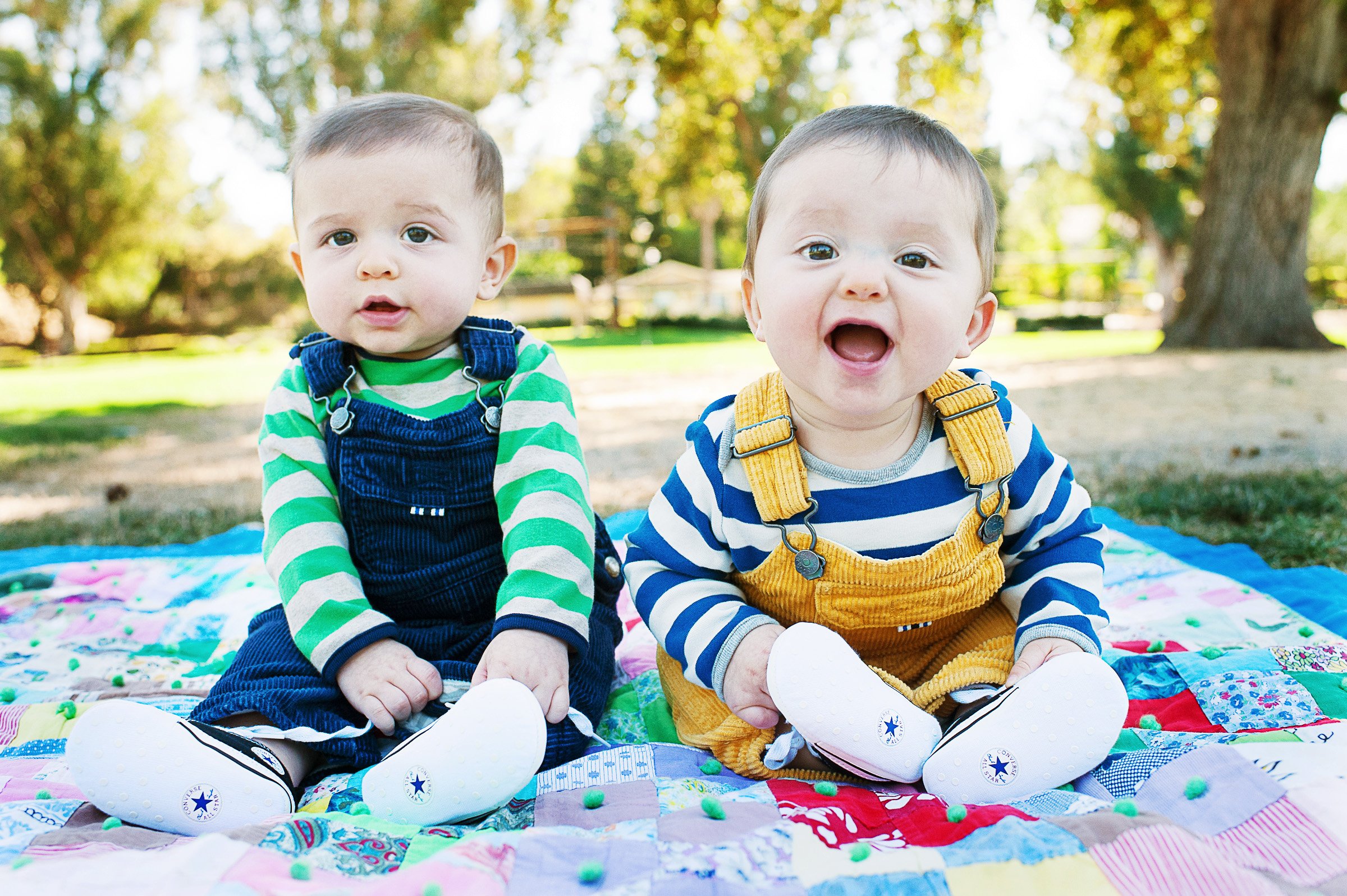 Two babies sitting on a colorful quilt outdoors, dressed in striped shirts and overalls, one with green stripes and denim overalls, the other with blue stripes and yellow overalls.