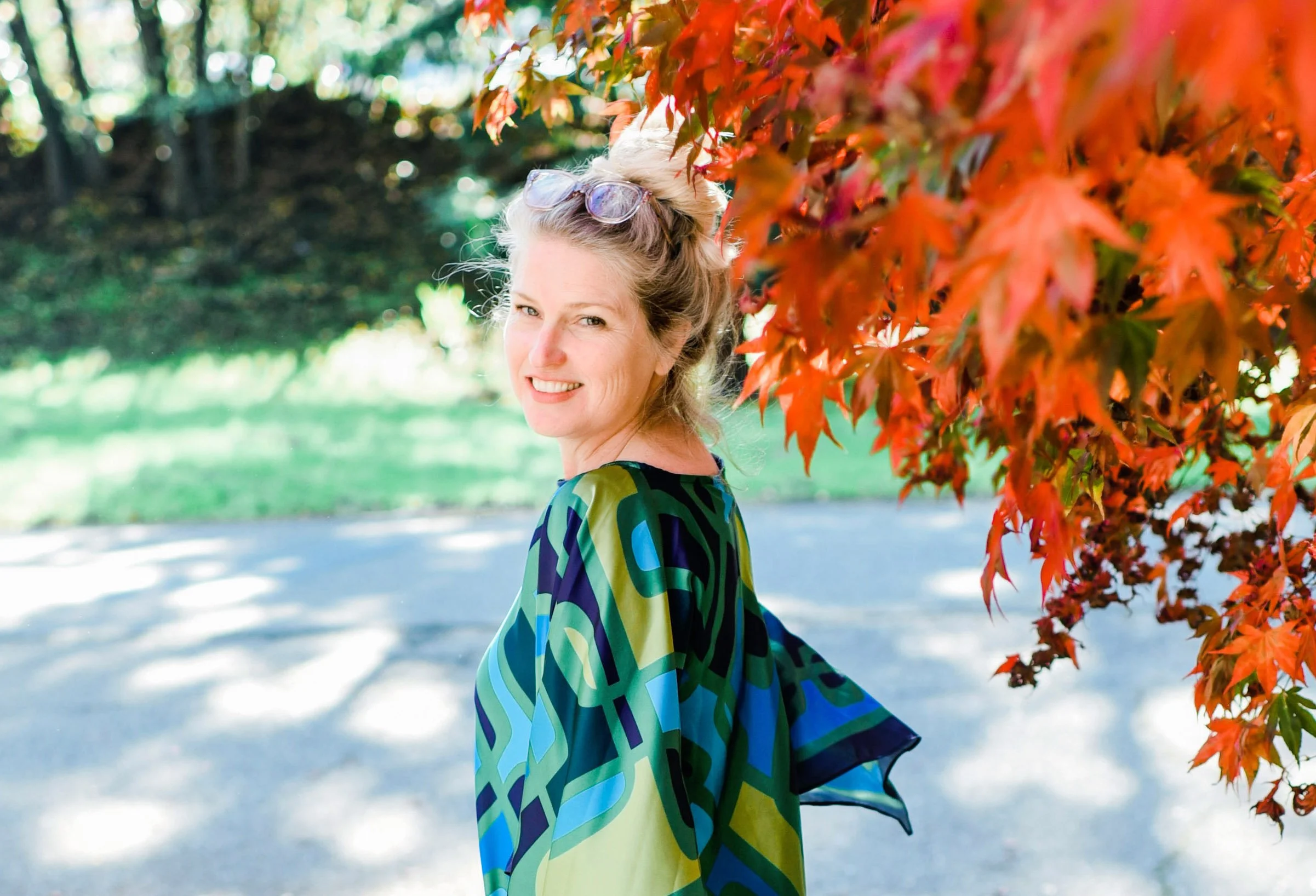 Woman smiling beside tree with red leaves, wearing colorful patterned top outdoors in a park.