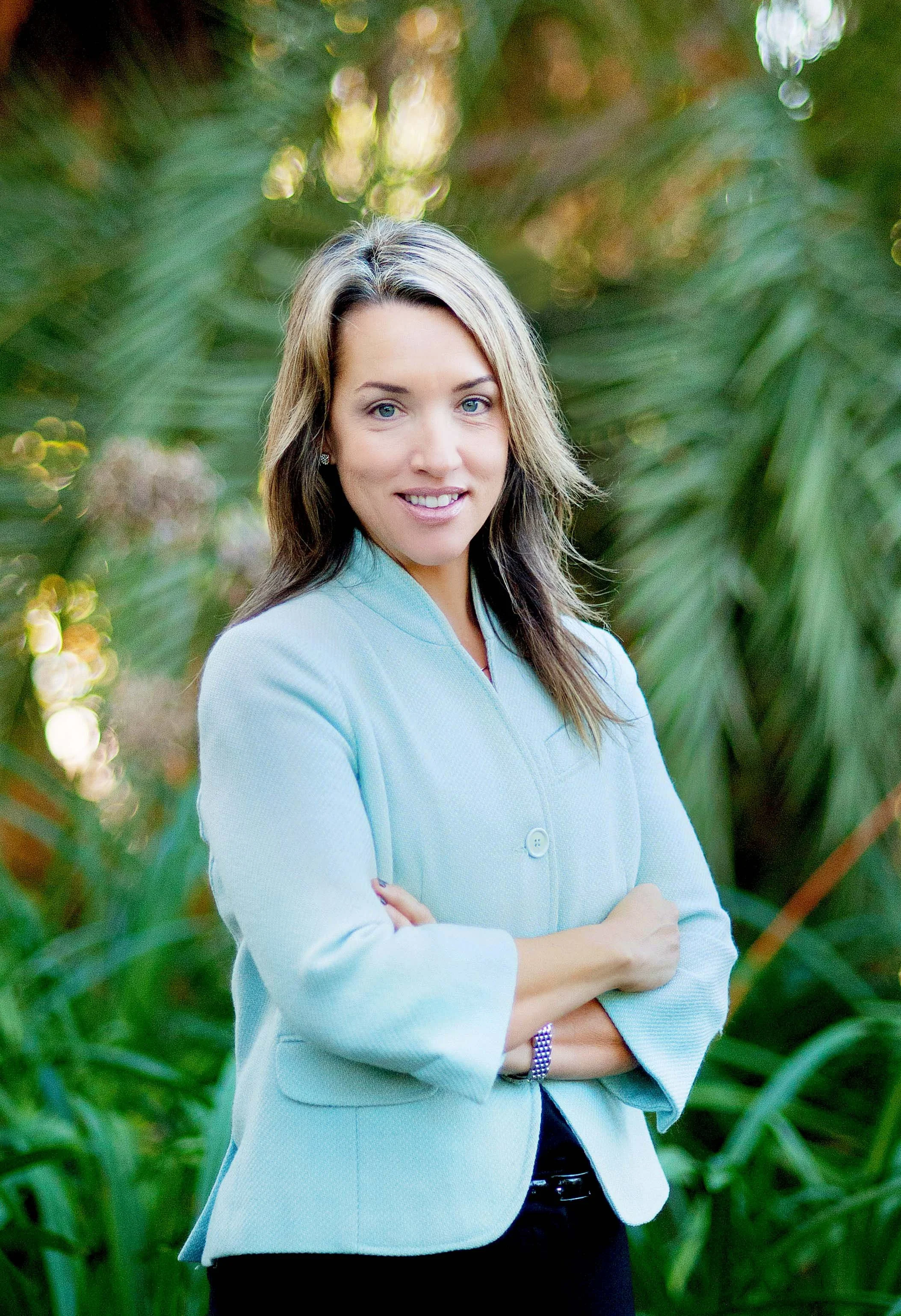 Woman in light blue jacket standing in front of green foliage.