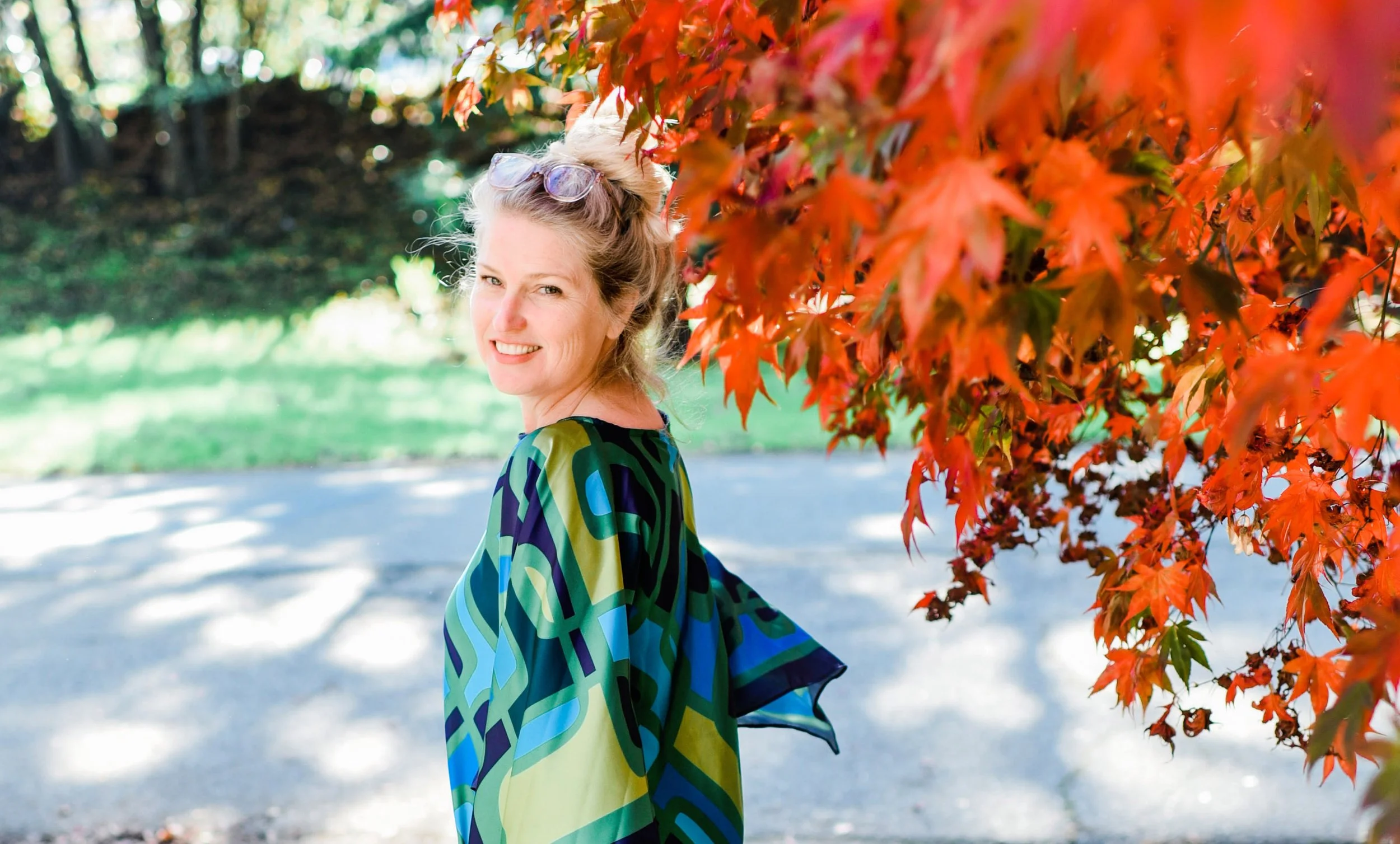 A woman with sunglasses on her head smiling outdoors next to red autumn leaves in a park.