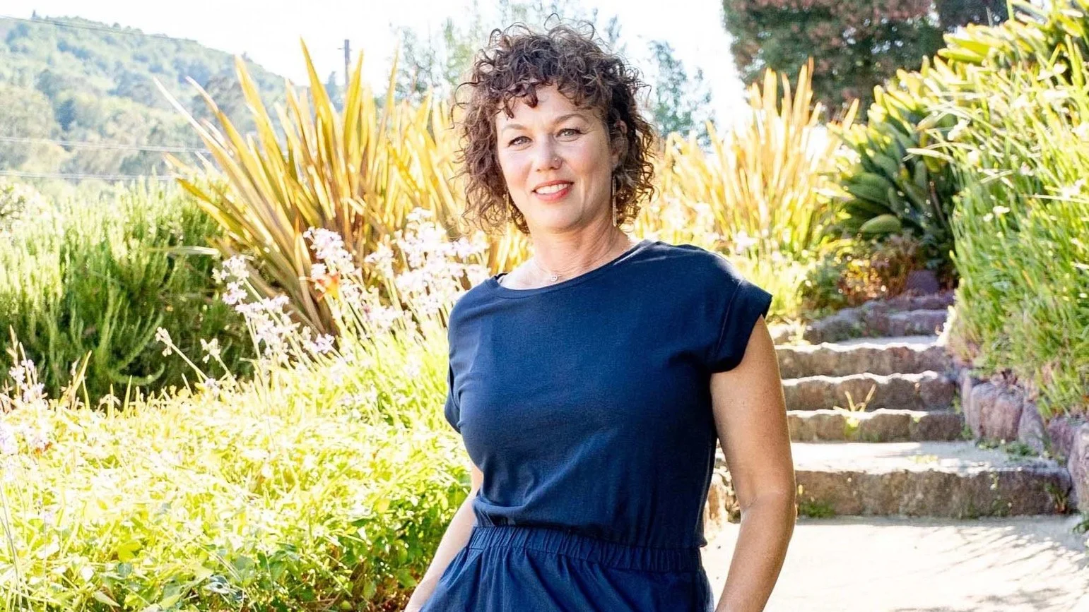 A woman with curly hair and light skin, wearing a navy blue short-sleeve dress, standing outdoors on a sunny day with a garden and stone steps in the background.