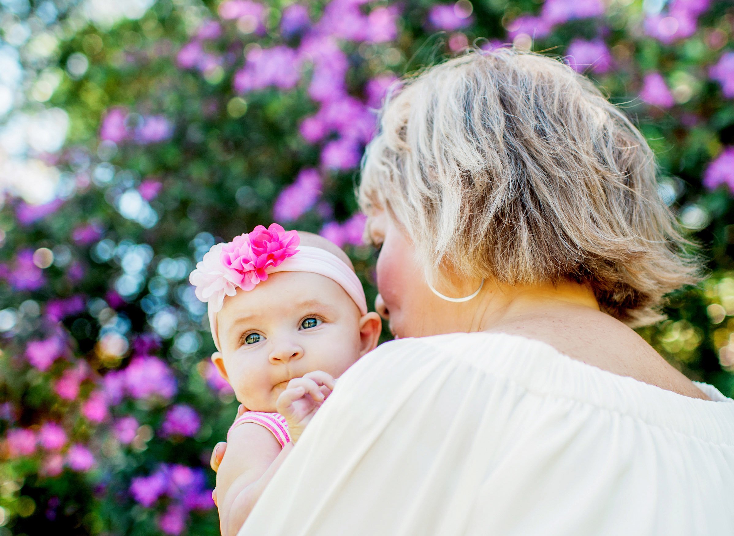 A woman holding a baby wearing a pink headband with flowers, set against a blurred background with purple flowers.