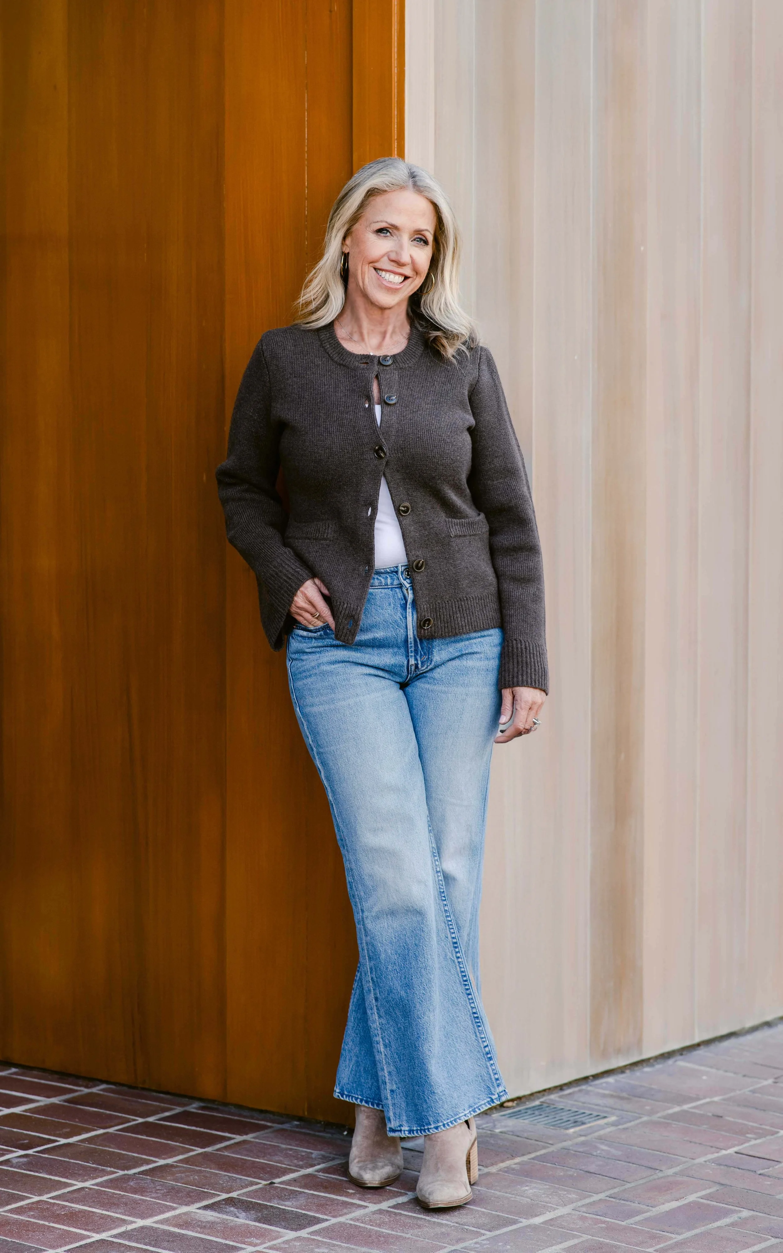A smiling woman with blonde hair standing with one hand in her pocket, wearing a dark brown cardigan, light blue jeans, and taupe ankle boots, leaning against a wooden wall.