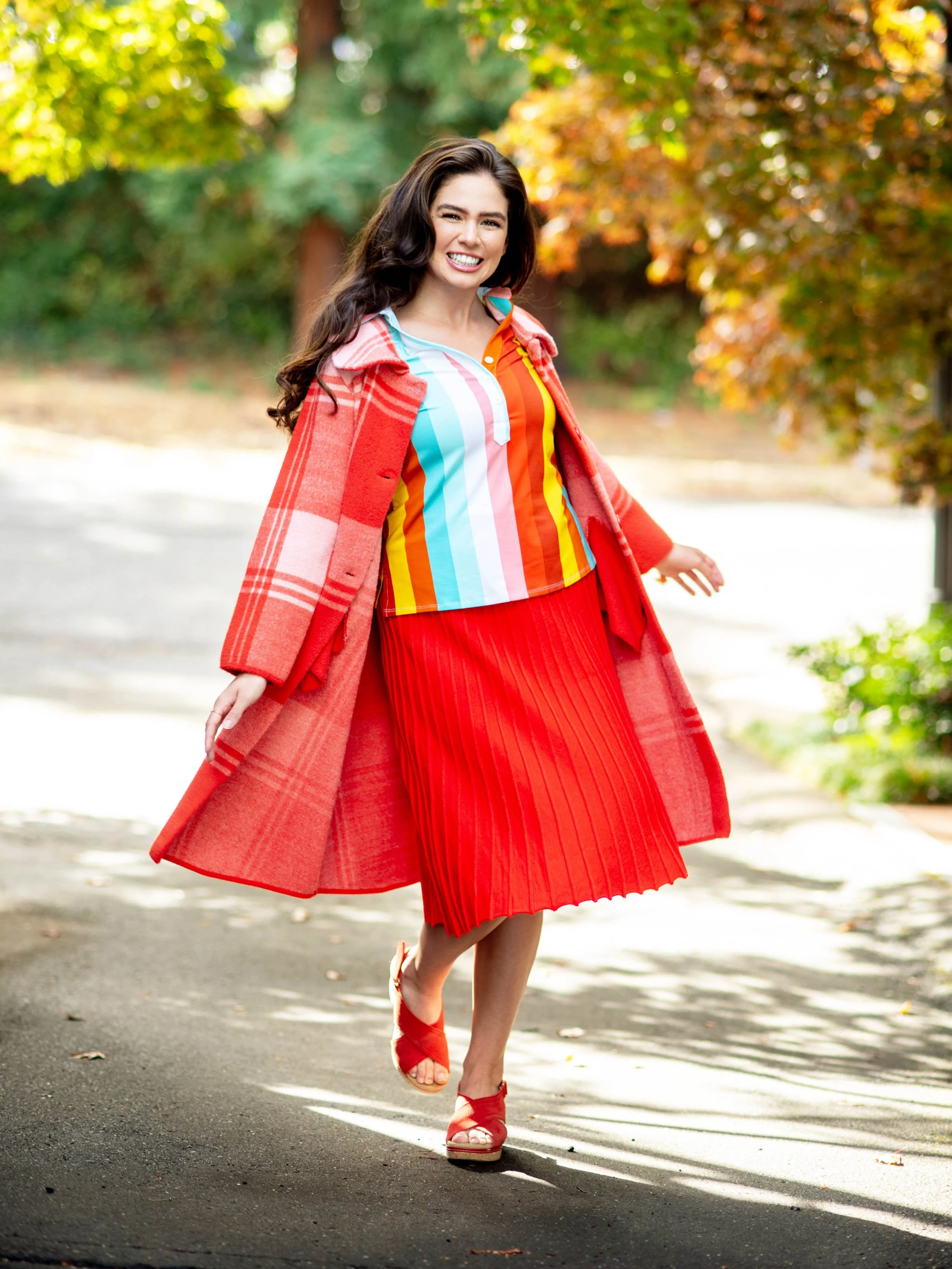 A woman with long dark wavy hair smiling and walking outdoors on a sunny day, wearing a colorful striped top, a red pleated skirt, red heels, and a red plaid coat, with trees and fall foliage in the background.